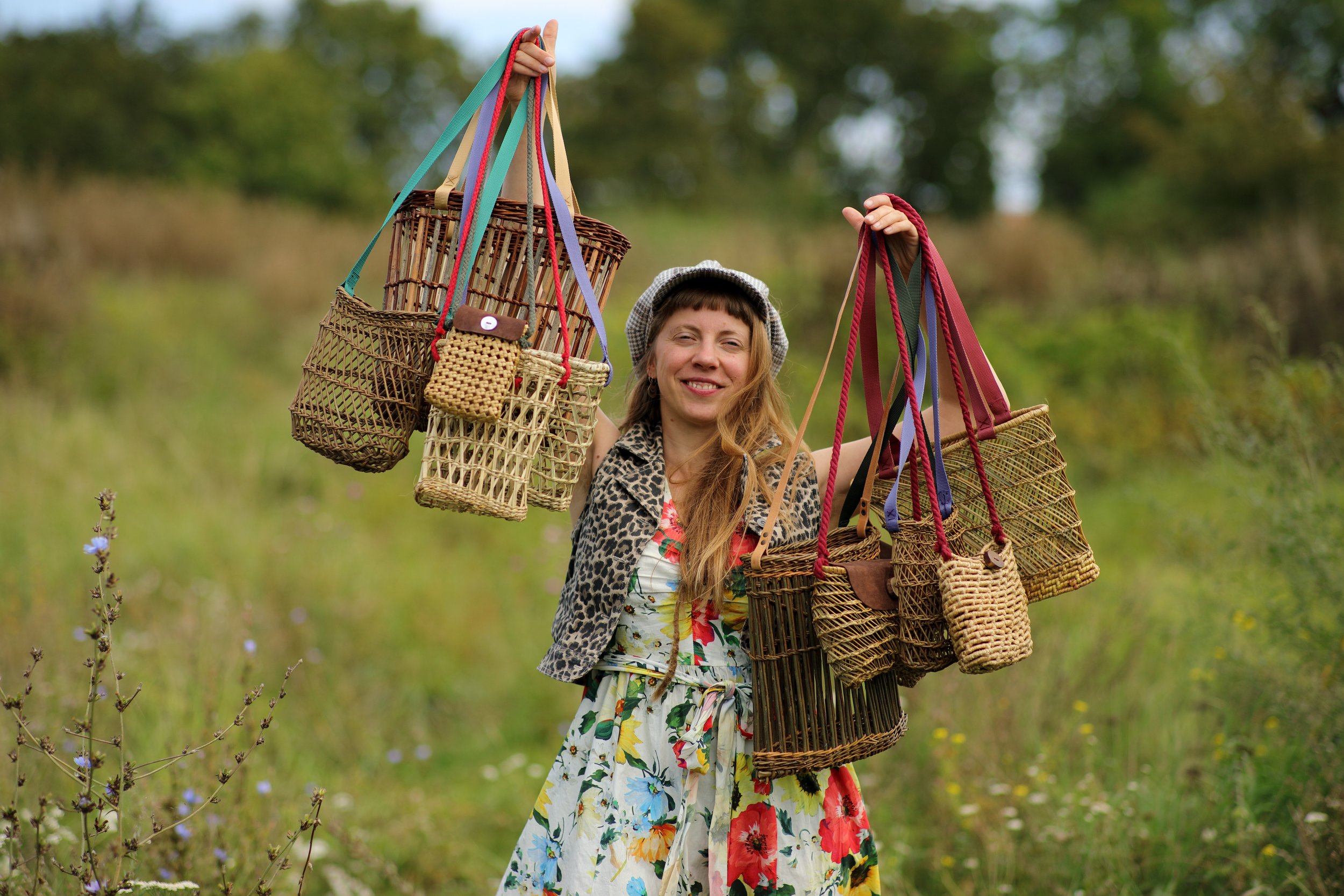 Mujer sonriendo, sosteniendo varias bolsas de mimbre y cestas en un campo de hierba y flores, con árboles en el fondo.
