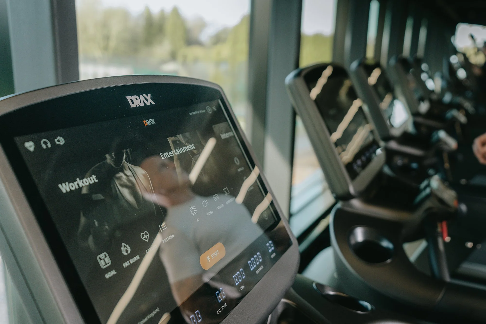 Close-up of the touchscreen on Drax cardio machine displaying workout options, with multiple cardio machines in the background.