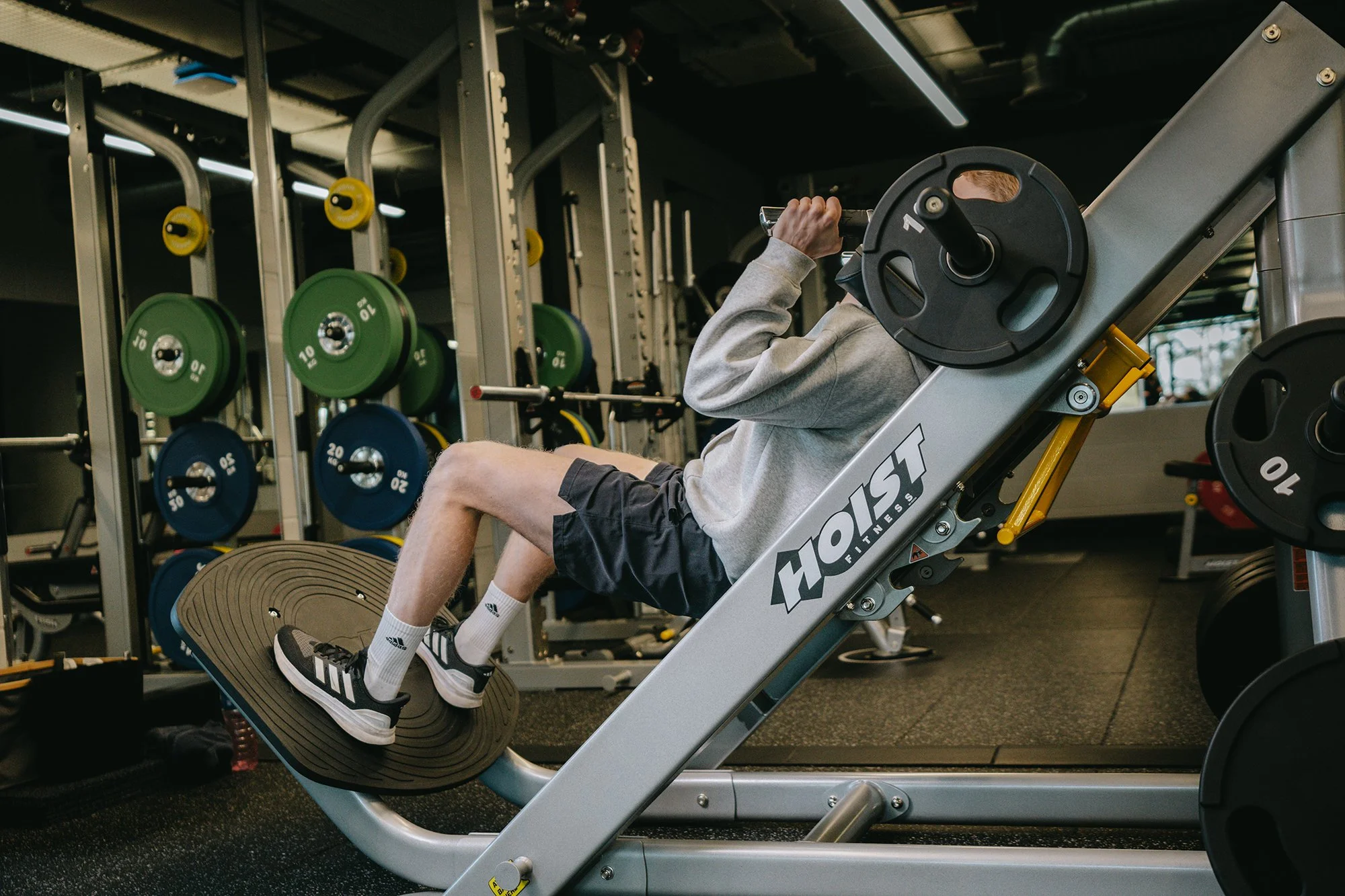 A person wearing a gray hoodie, black shorts on a Hoist leg press machine in a gym, with weight plates visible on a rack and other gym equipment in the background.