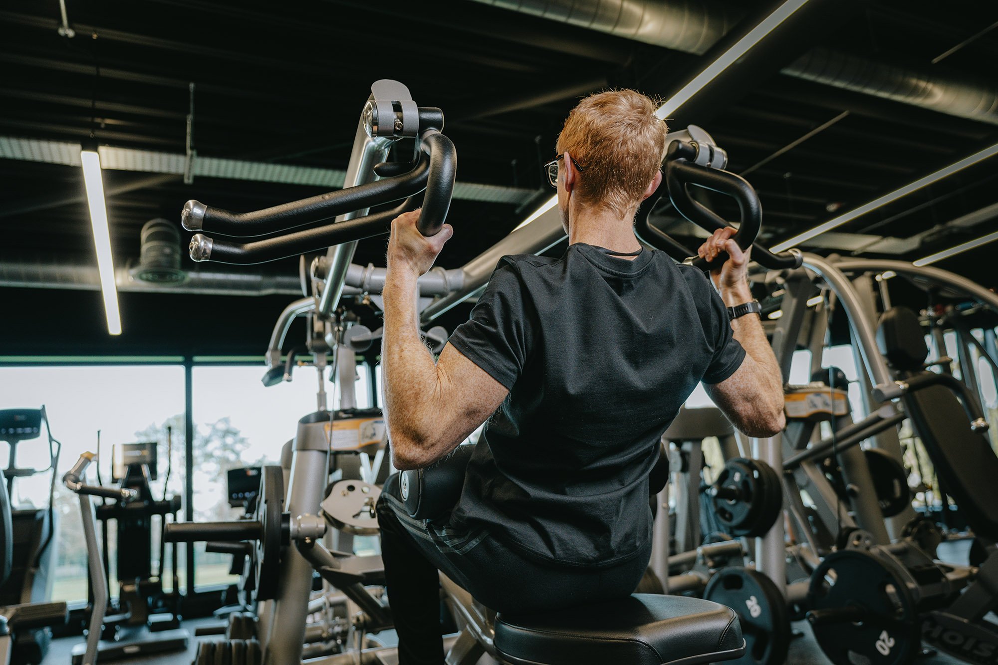 A man with red hair and glasses is using a lat pulldown machine with various exercise equipment around him.