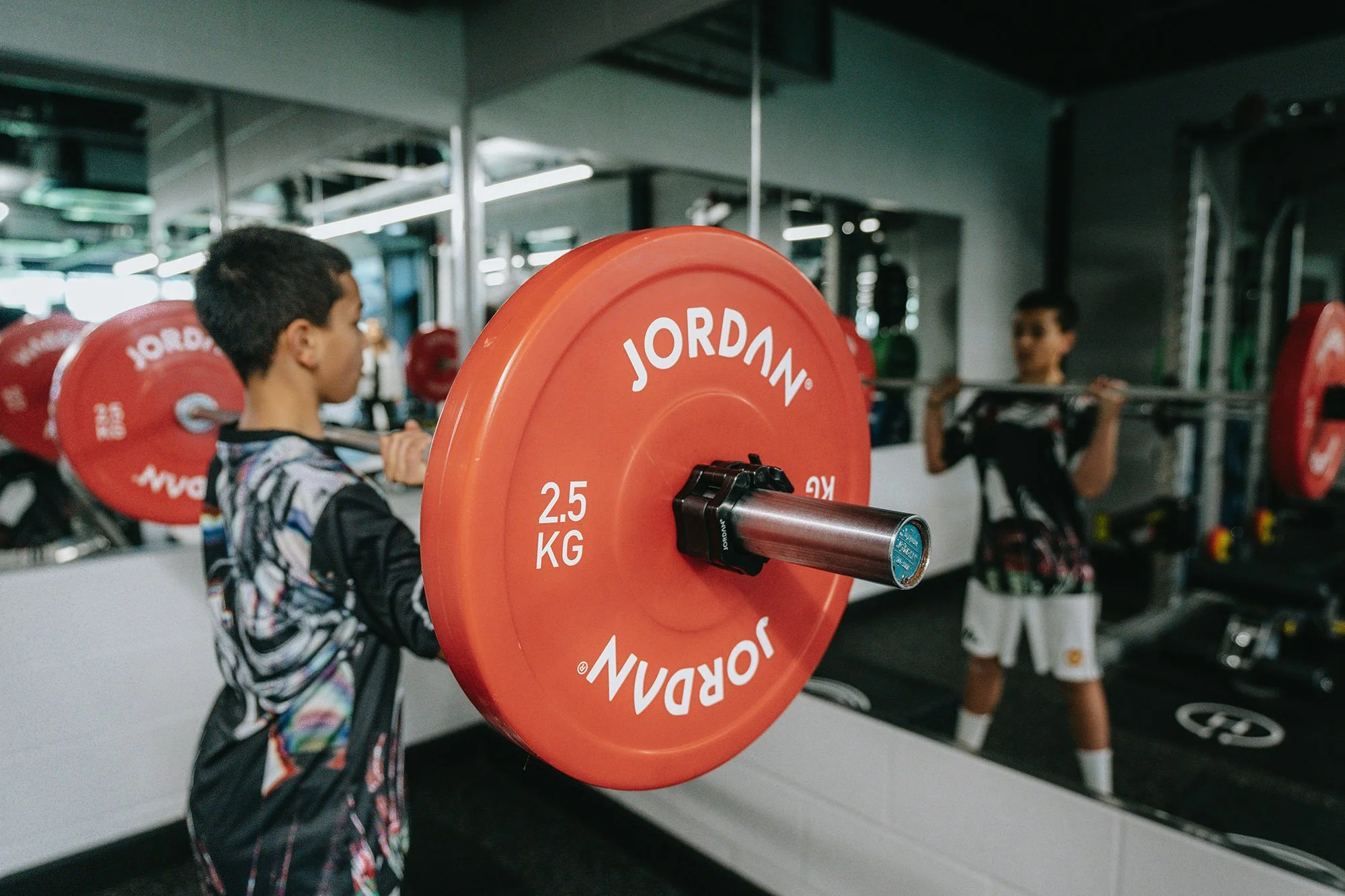 Person lifting a barbell with red weight plates - other gym equipment and mirrors are visible.