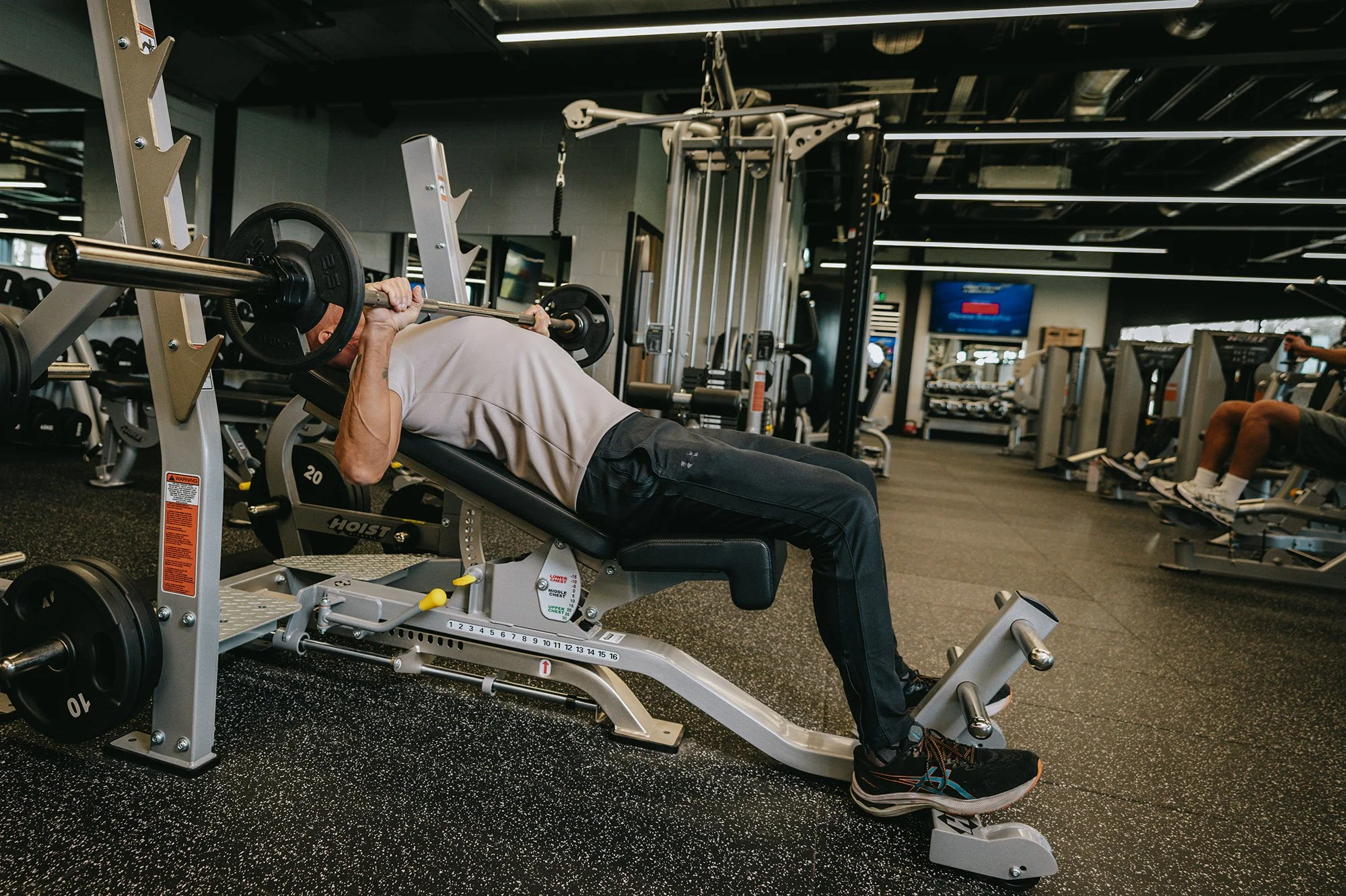 Person lying on a weight bench lifting a barbell at a gym, surrounded by workout equipment.