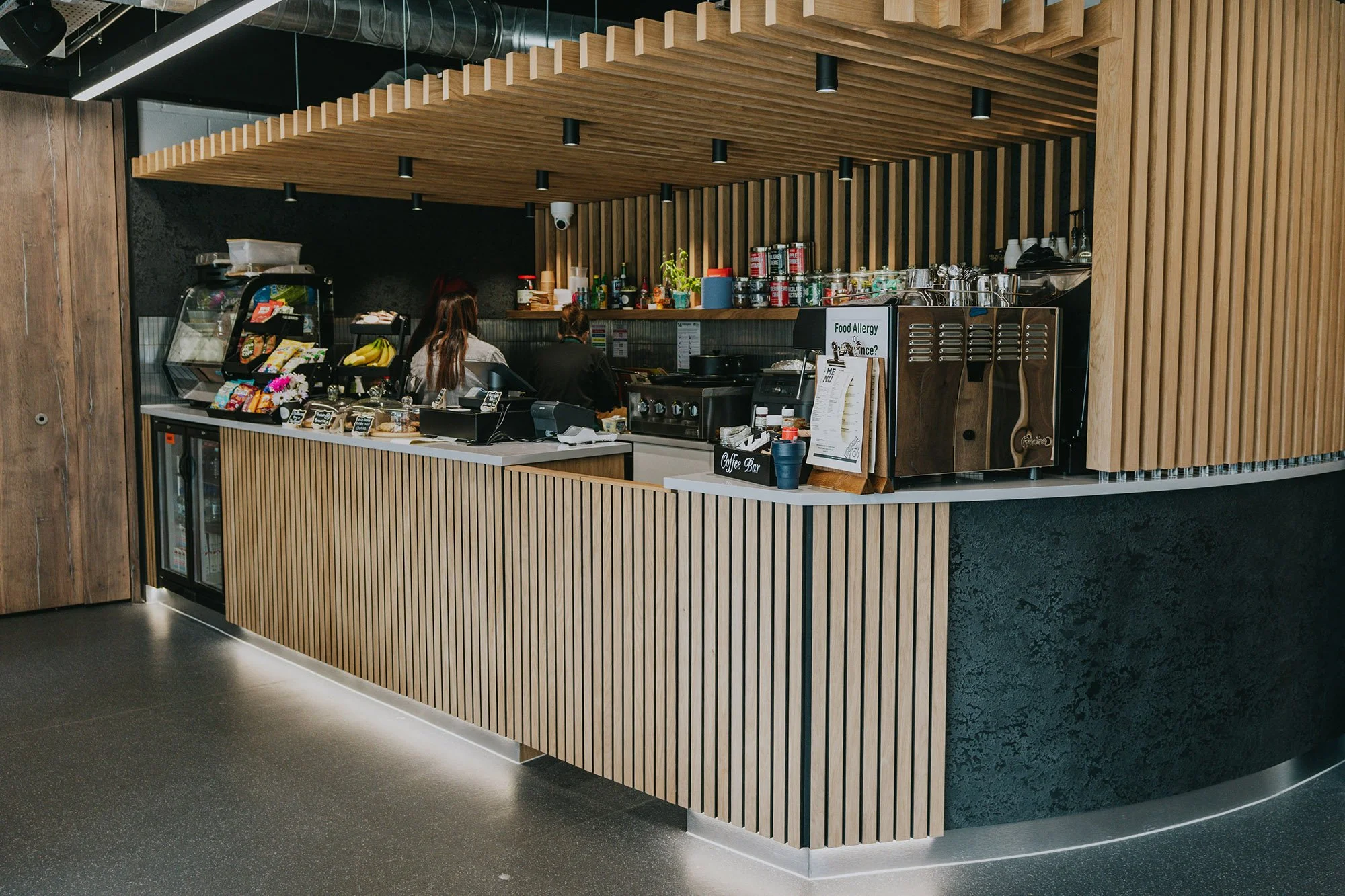 Cup of Joy wellness café counter with a wooden slatted front, coffee equipment, and a menu sign, with two people working behind the counter.
