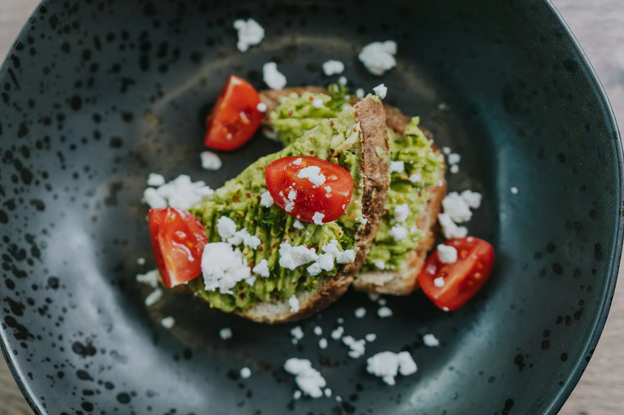 A black bowl containing guacamole topped with cherry tomato halves, crumbled white cheese, and a meat or plant-based topping, garnished with chopped green herbs.