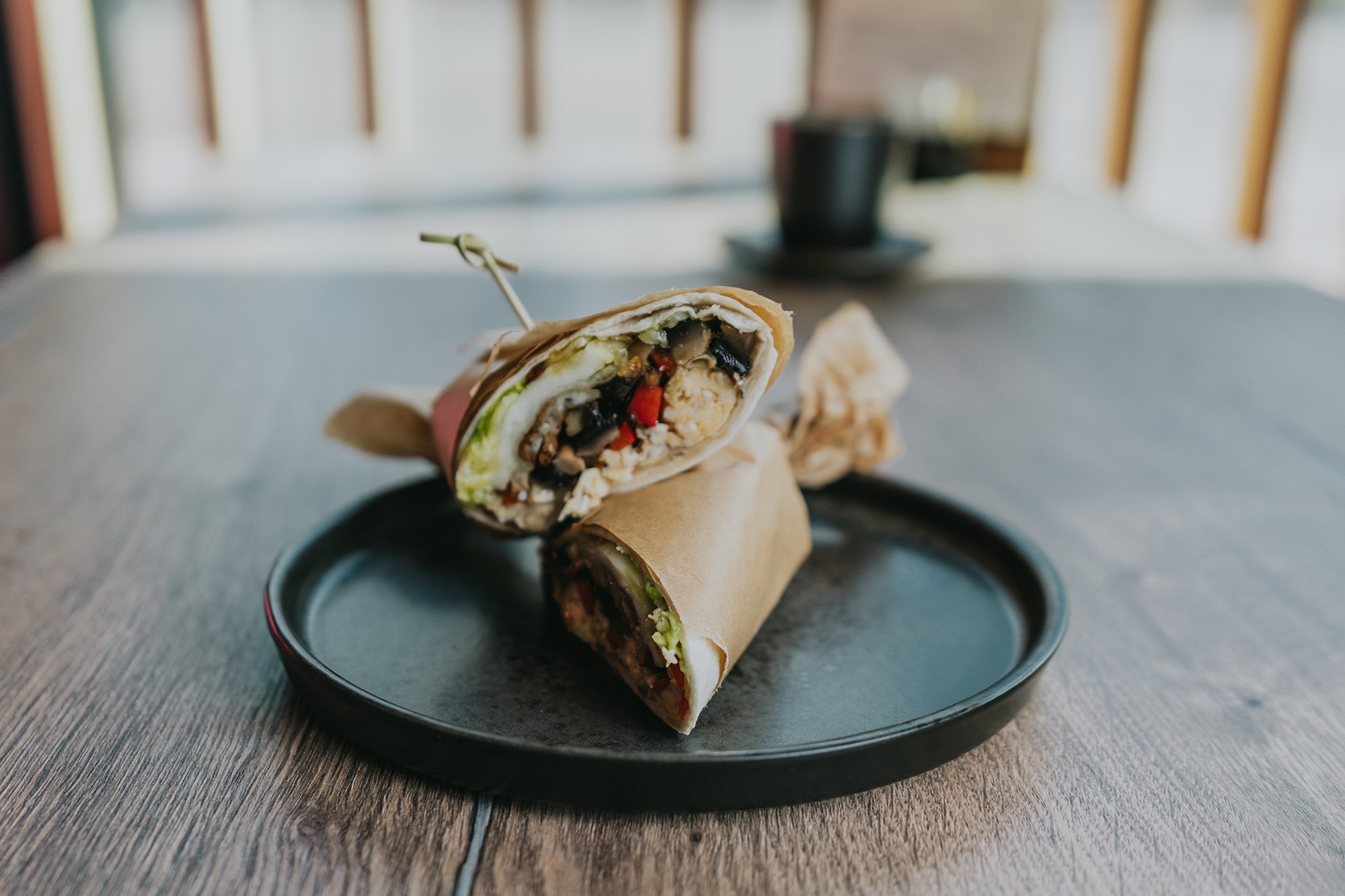 A burrito cut in half on a black plate, showing vegetables, beans, and rice inside. The plate is on a wooden table, with a blurred background of a railing and a cup.
