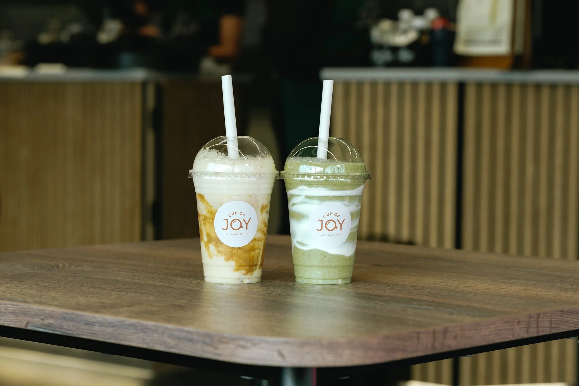 Two blended drinks in clear plastic cups with straws on a wooden table, each with a label that reads 'A Cup of Joy at Hamiltons'.