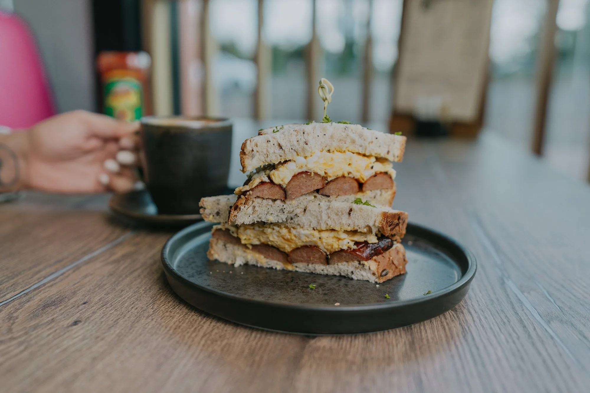 Stacked breakfast sandwich with sausage patties, scrambled eggs, and a toasted bun on a black plate at a wooden table, with a black coffee mug and blurred background.
