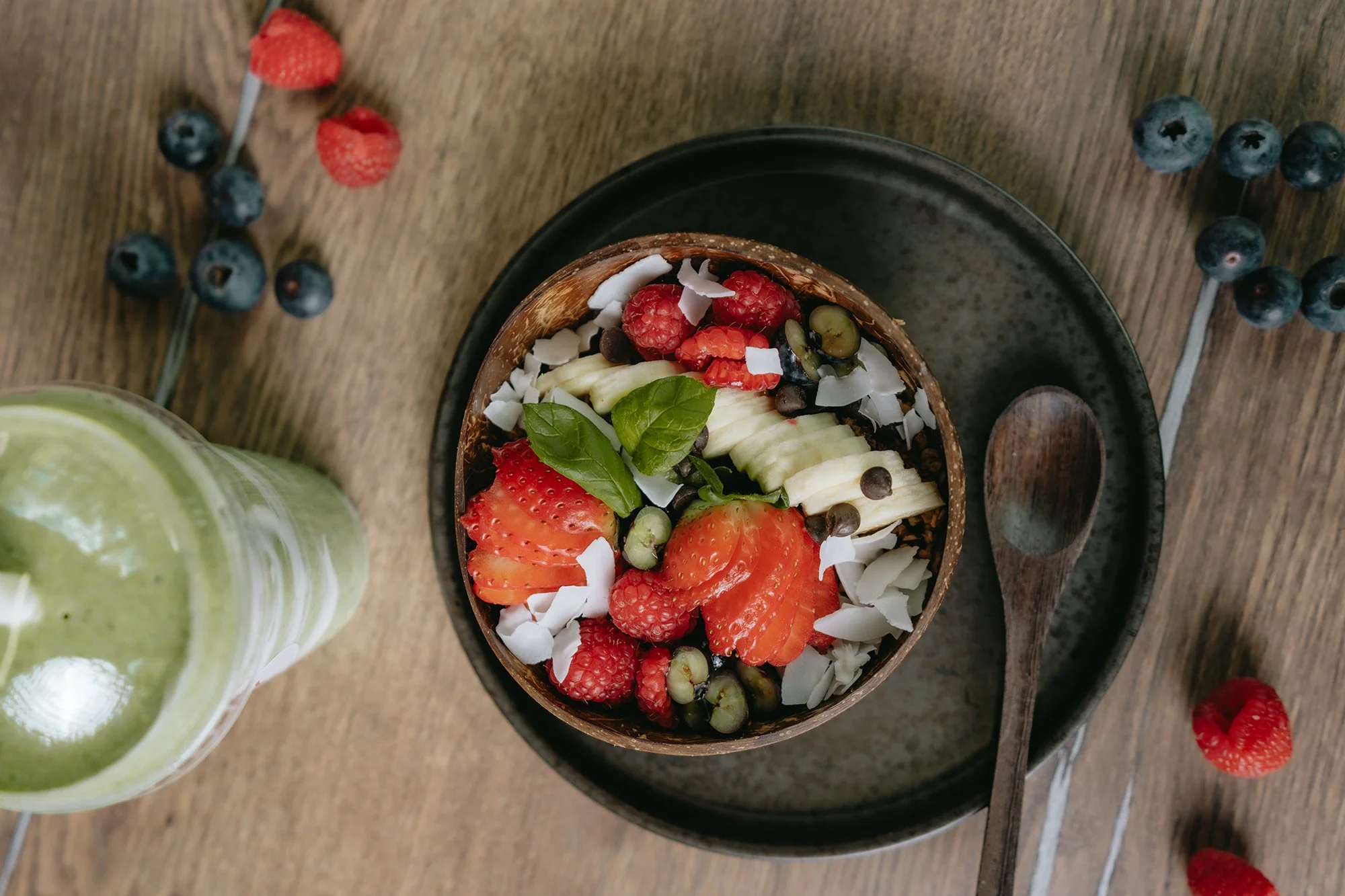 A bowl of smoothie topped with fresh strawberries, raspberries, banana slices, blueberries, coconut flakes, chocolate chips, and basil leaves, placed on a wooden table with scattered blueberries and raspberries around.