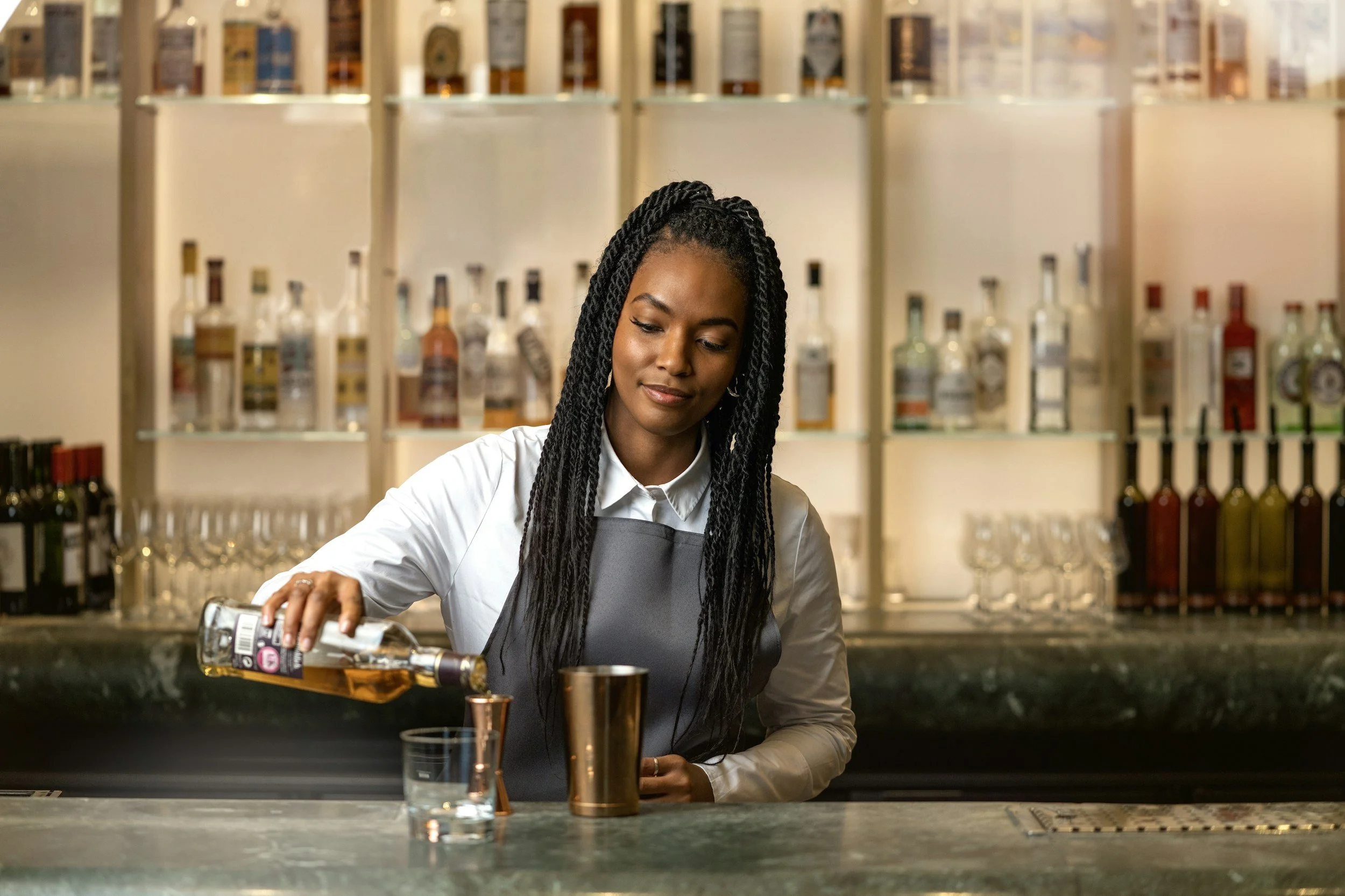 A woman with long black braided hair pouring whiskey into a glass at a bar with bottles in the background.