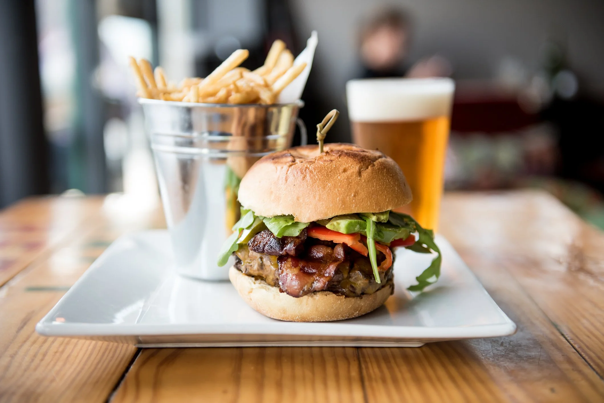 A burger with lettuce, tomato, and bacon, served on a bun with a skewer, accompanied by French fries in a metal cup and a glass of beer, all on a white plate on a wooden table.