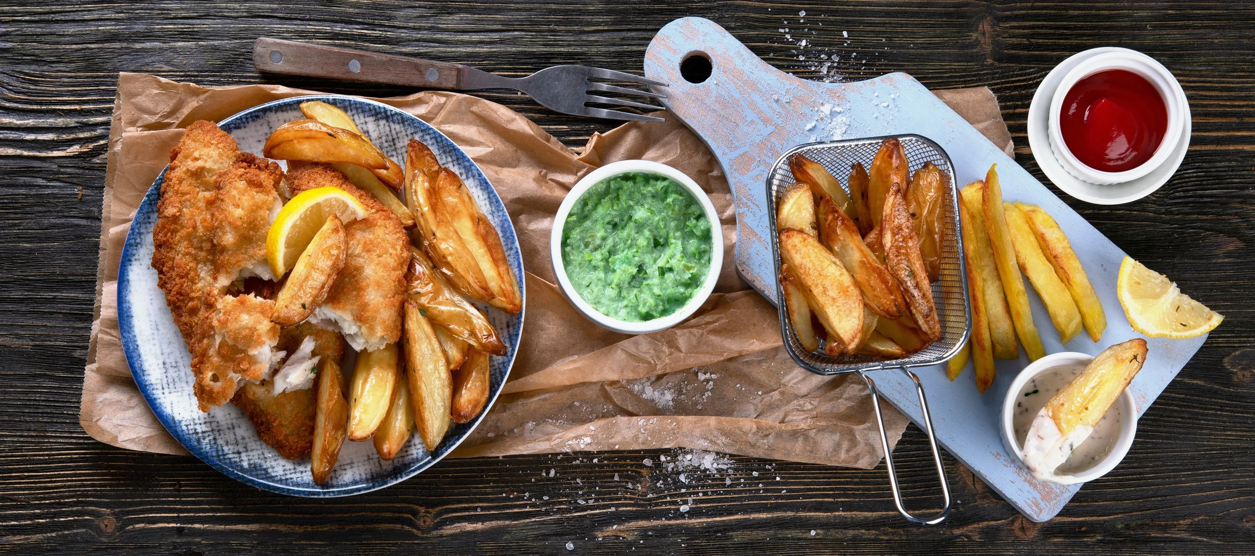 A top-down view of a meal including fried fish with lemon, potato wedges, tartar sauce with lemon, coleslaw, ketchup, and lemon wedges on a rustic wooden table.