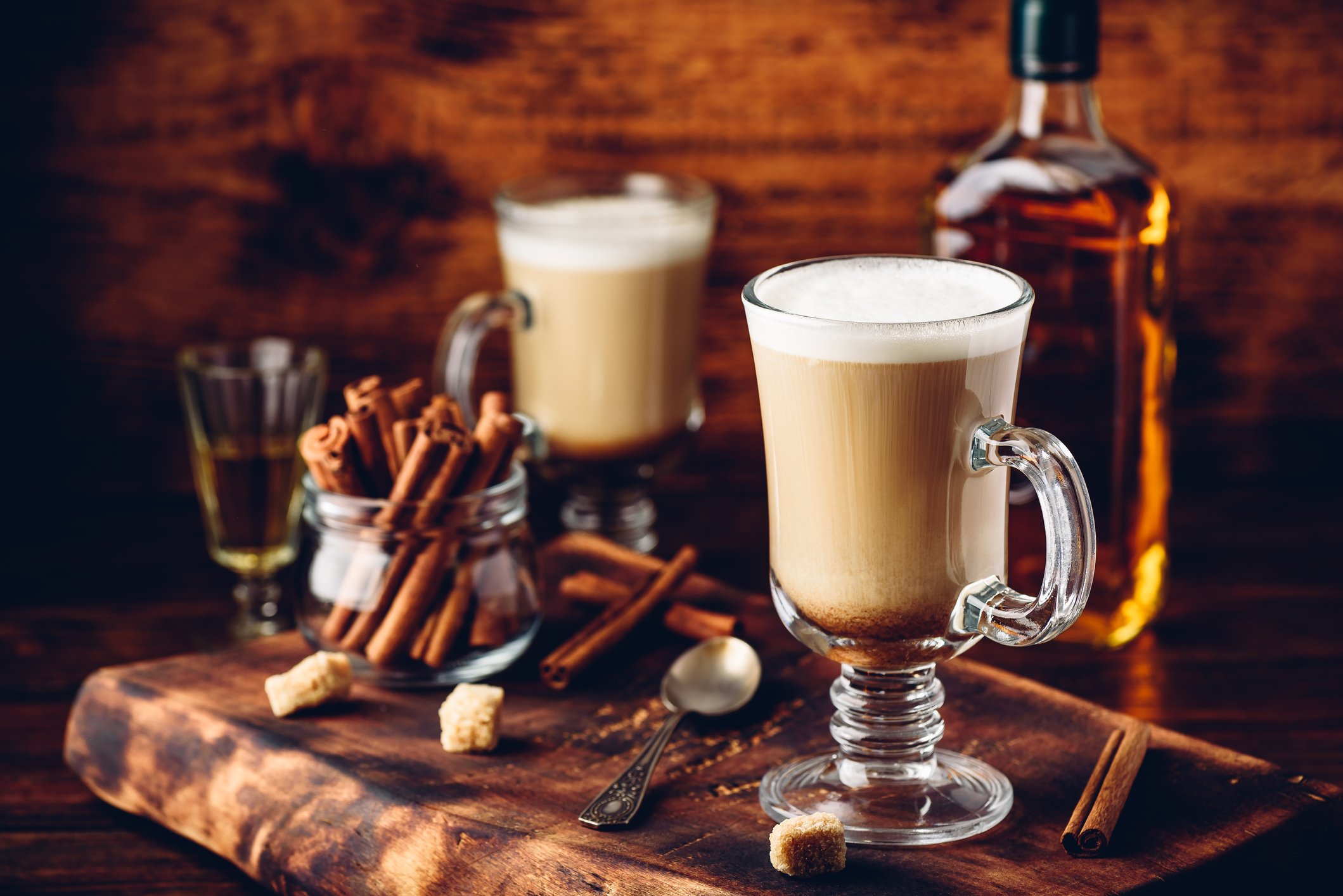 Two Irish coffee glasses with whipped cream on top, a bottle of whiskey, cinnamon sticks in a jar, sugar cubes, and a teaspoon on a wooden surface.