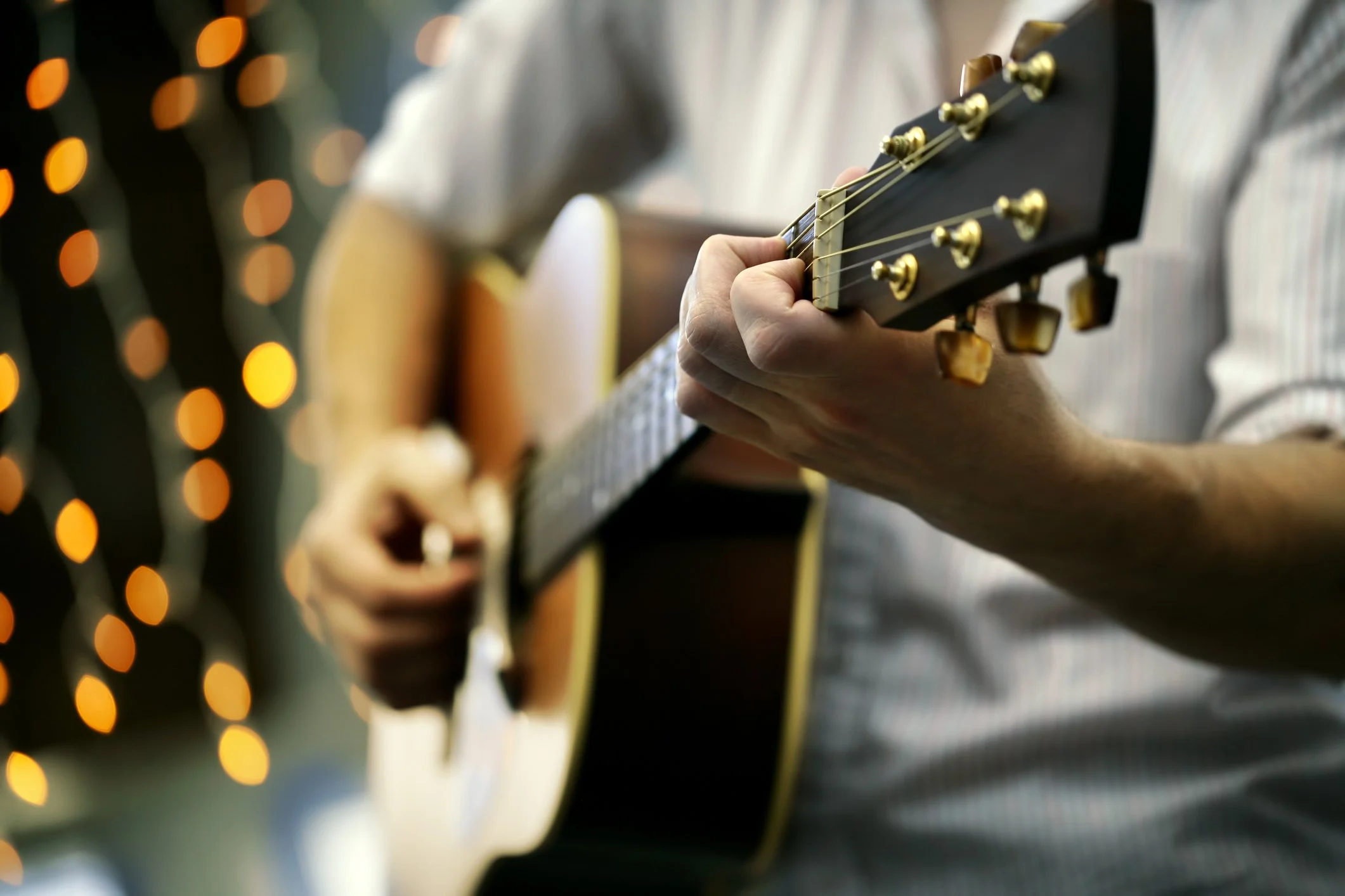 A person playing an acoustic guitar, focusing on their hand on the fretboard, with blurred background lights.