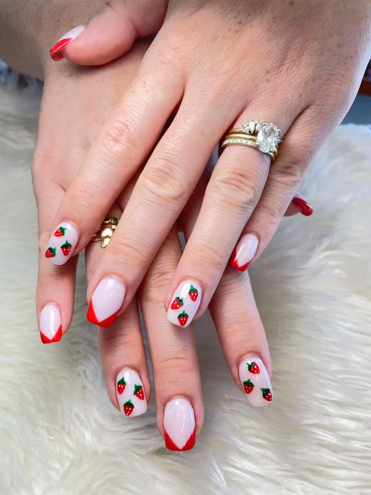 Close-up of a person's hands with decorated nails featuring white and red strawberry-themed nail art, resting on a soft, fluffy white surface. The person is wearing multiple rings, including a large oval diamond engagement ring and a gold band with small diamonds.
