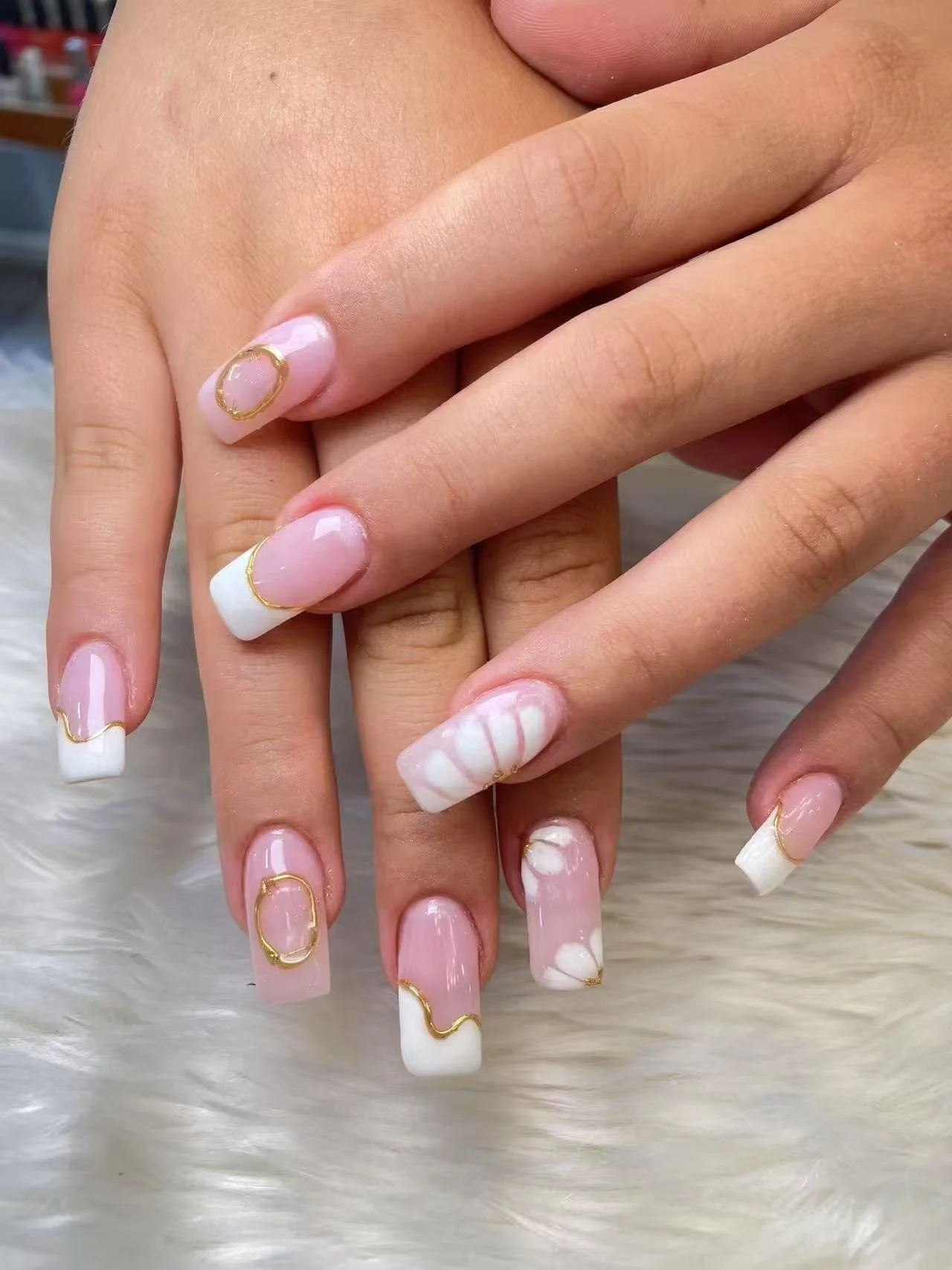 Close-up of hands with manicured nails featuring white and pink French tips with gold accents and marble designs.