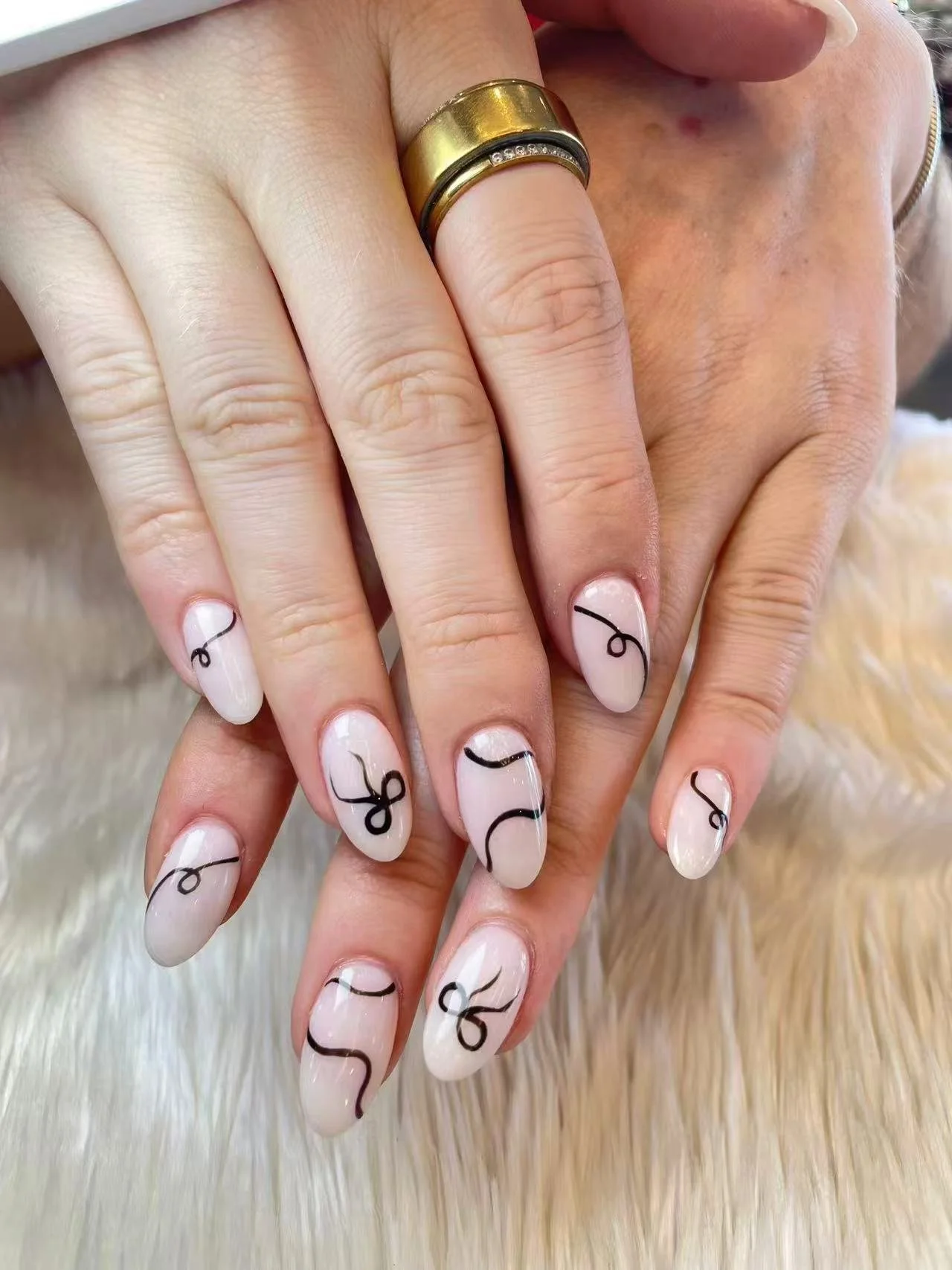 Close-up of hands with manicured nails painted in a light color with black, curly designs. One hand has a gold ring and another has a ring with small stones.