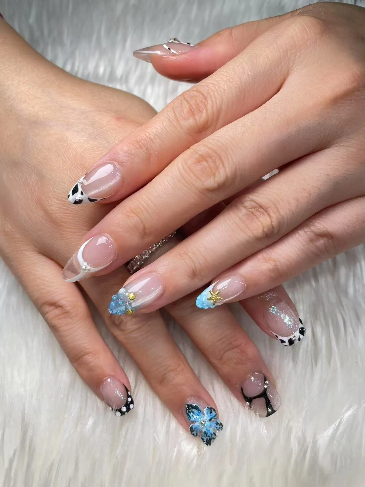 Close-up of hands with decorated acrylic nails featuring beach-themed designs, including flowers, starfish, cow print, and glitter accents on a neutral background.