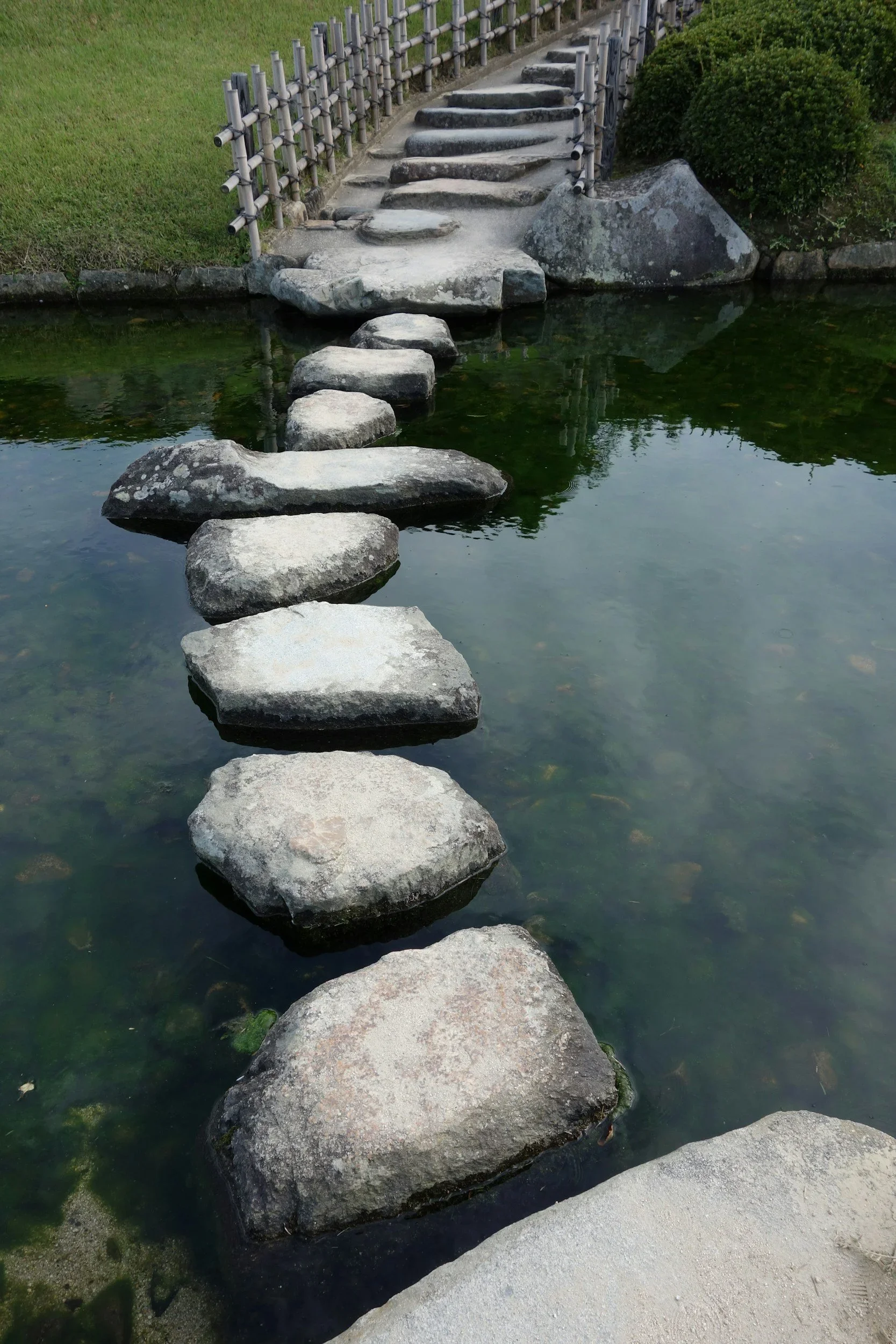 Stone pathway crossing a pond with stepping stones, leading to a small bridge and garden area with bushes and a bamboo fence.