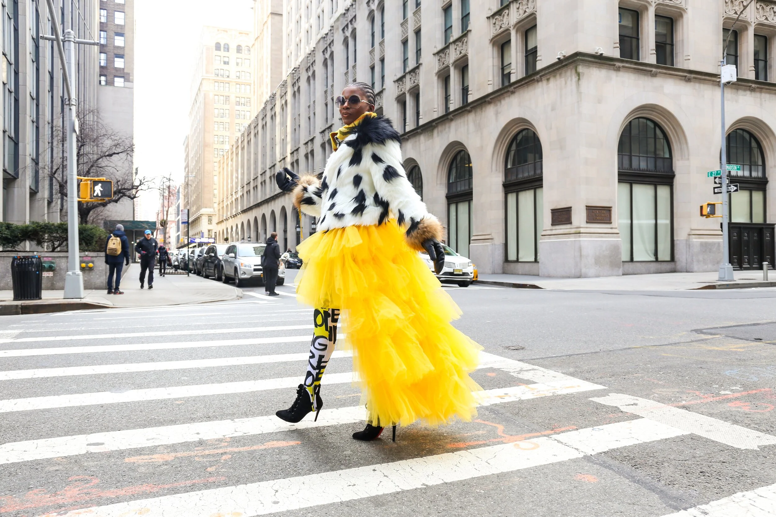 A woman in a yellow tulle skirt, black high heels, animal-print stockings, a black and white faux fur coat, yellow scarf, black gloves, and large sunglasses crossing a city street.