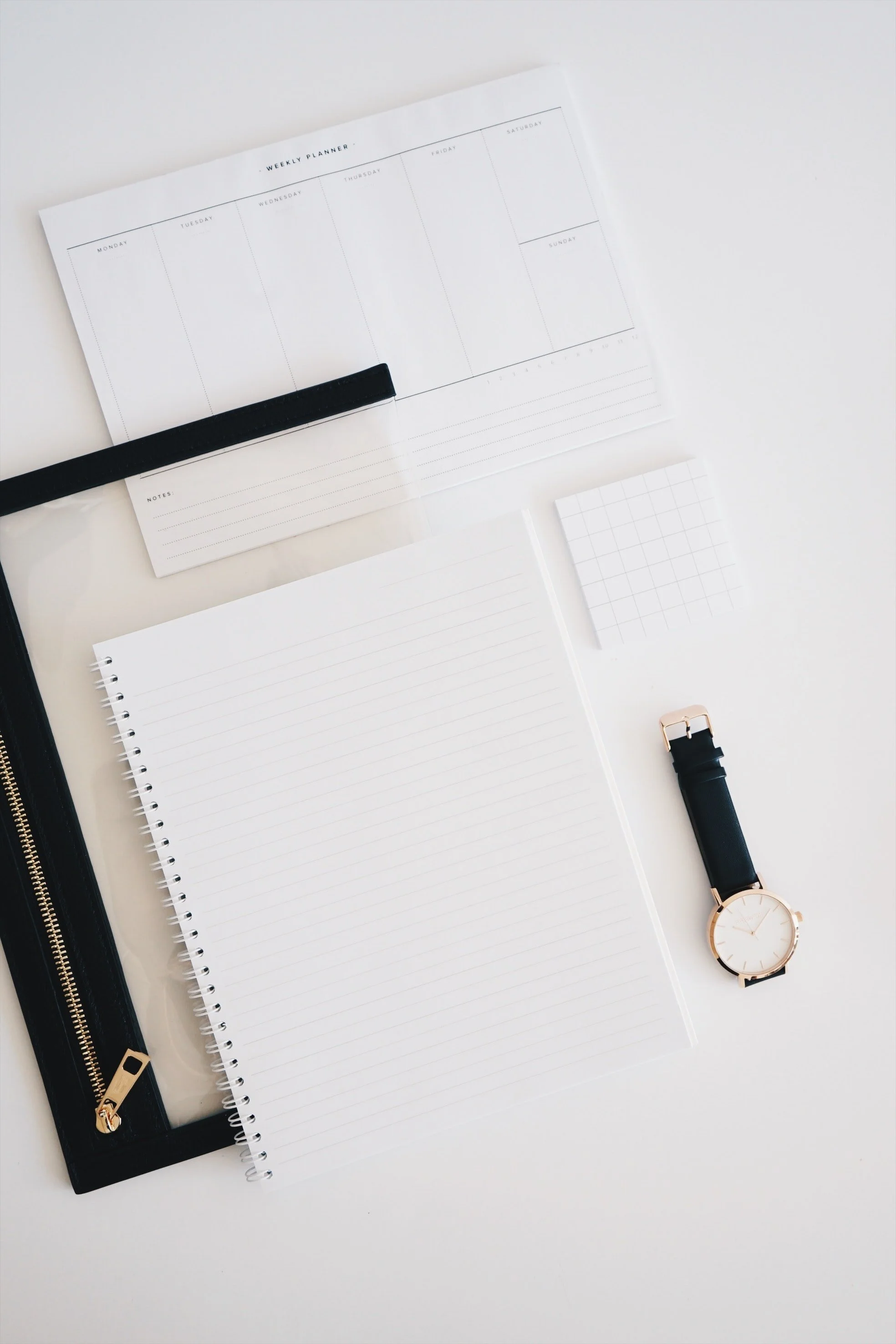 A flat lay of a white planner, spiral notebook, small grid paper, a black watch with gold accents, and a black zippered case on a white surface.