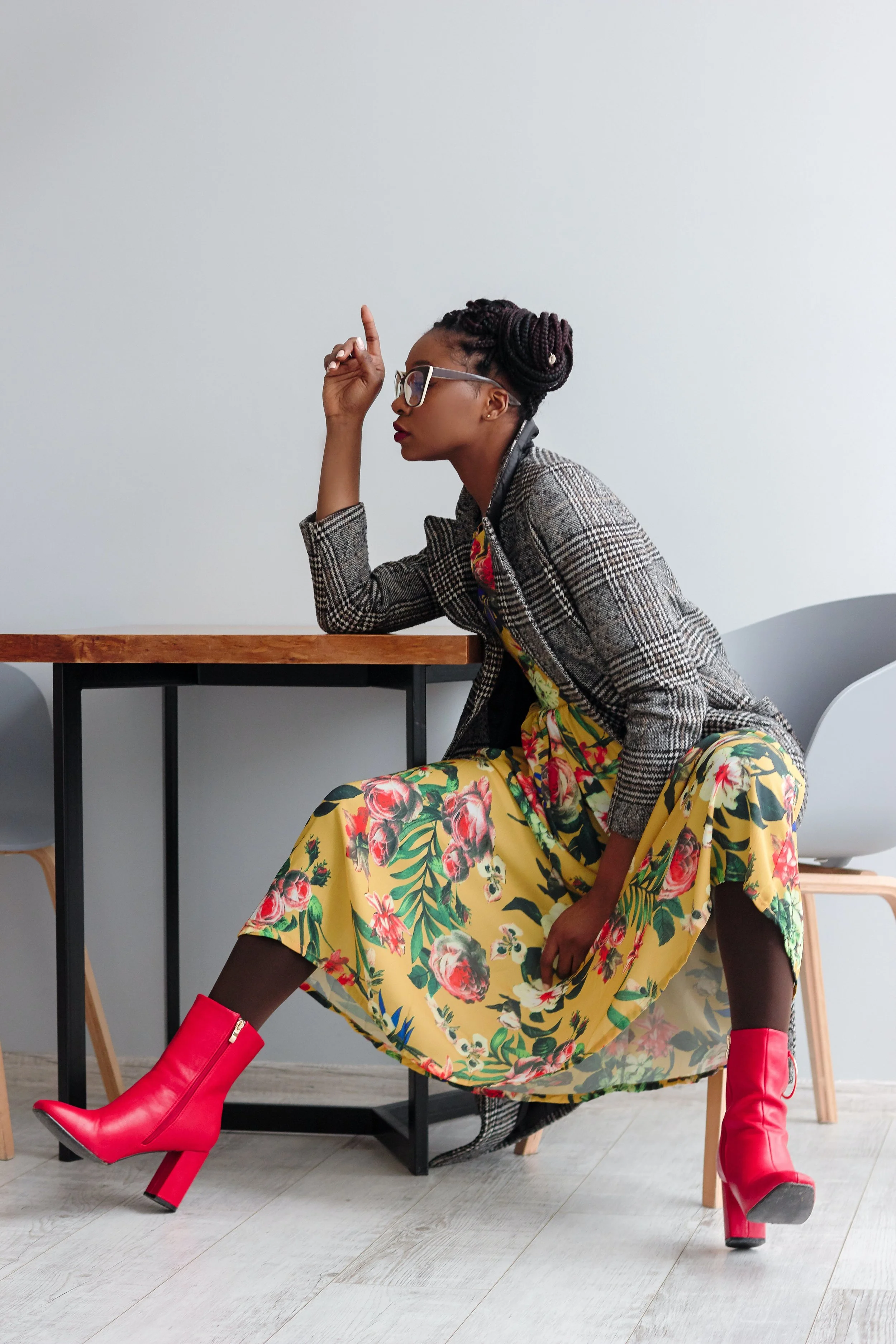 A woman sitting on a wooden chair with one foot on the ground and the other foot resting on the chair's crossbar. She is wearing red ankle boots, a floral yellow dress, a gray plaid blazer, and glasses. She has her hair styled in dreadlocks and is posed with her right hand raised, finger pointing upward, in front of a plain white wall.