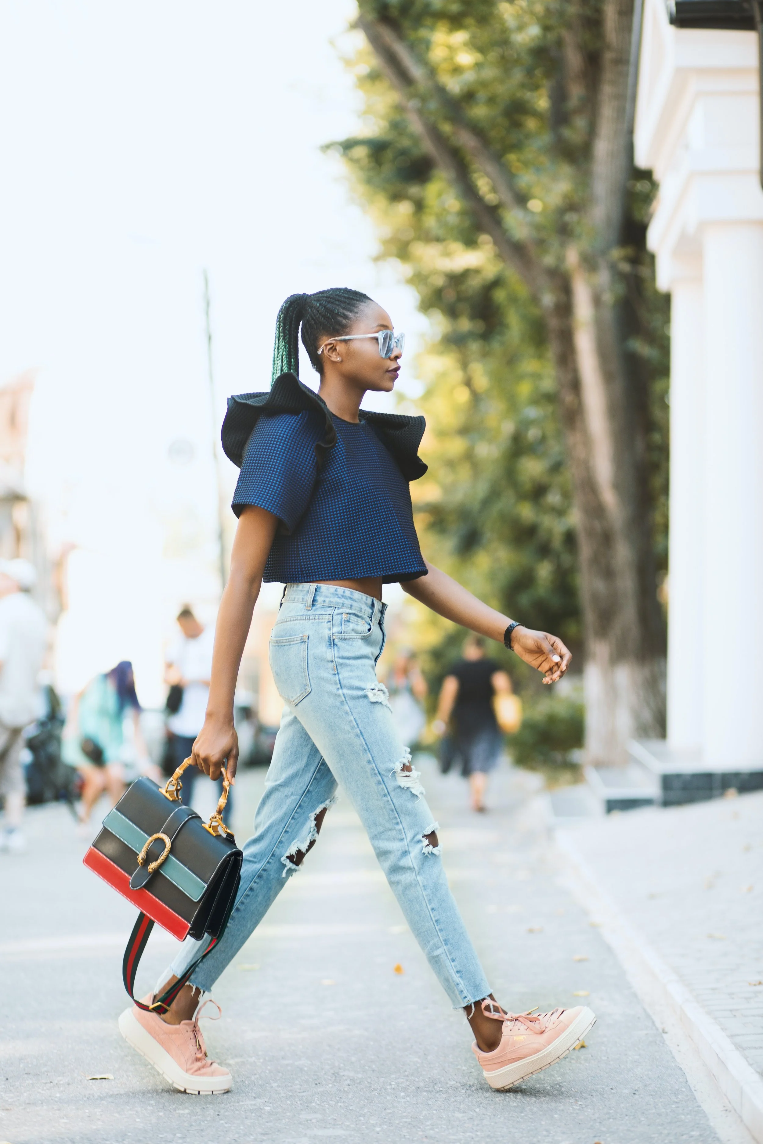 A young woman walking on a city street during daytime, wearing a blue crop top, ripped jeans, pink sneakers, sunglasses, and carrying a colorful handbag.