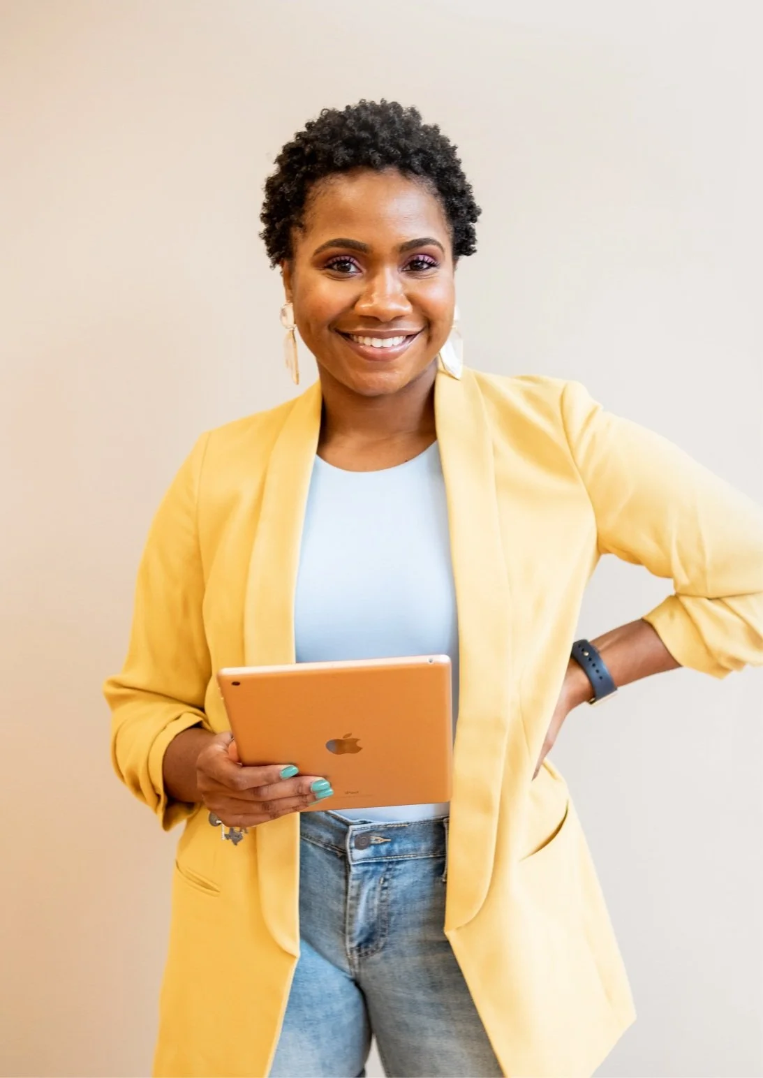 A woman with short curly hair, wearing a yellow blazer and light blue shirt, holding an orange tablet, standing against a plain off-white wall, smiling at the camera.