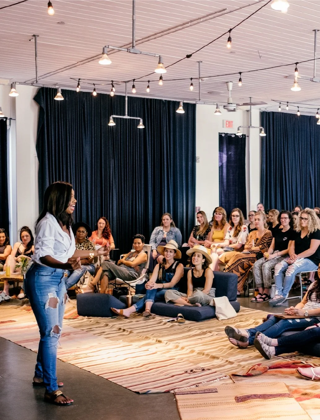 A woman standing and speaking to an audience of women in a room with black curtains and string lights.