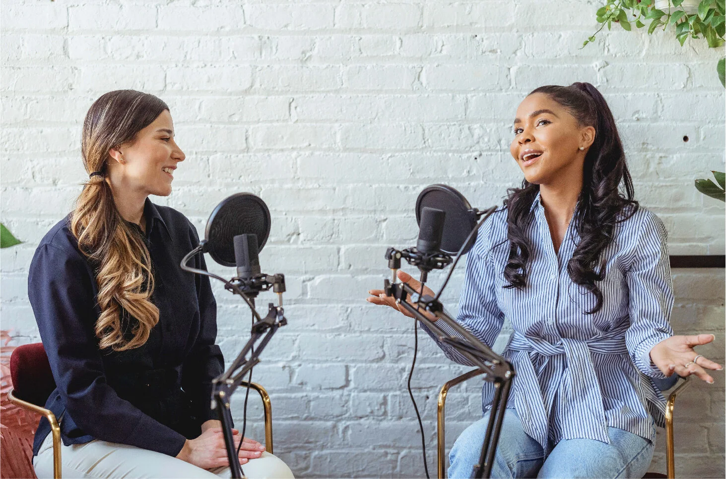 Two women having a conversation while recording a podcast, sitting on chairs with microphones in front of them, against a white brick wall.