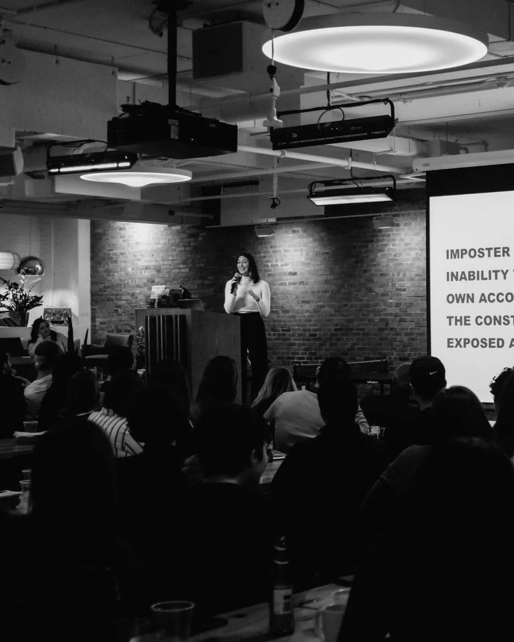 A woman on stage giving a presentation in a dimly lit room with an exposed brick wall. Audience members are seated facing her, and a large screen displays text.