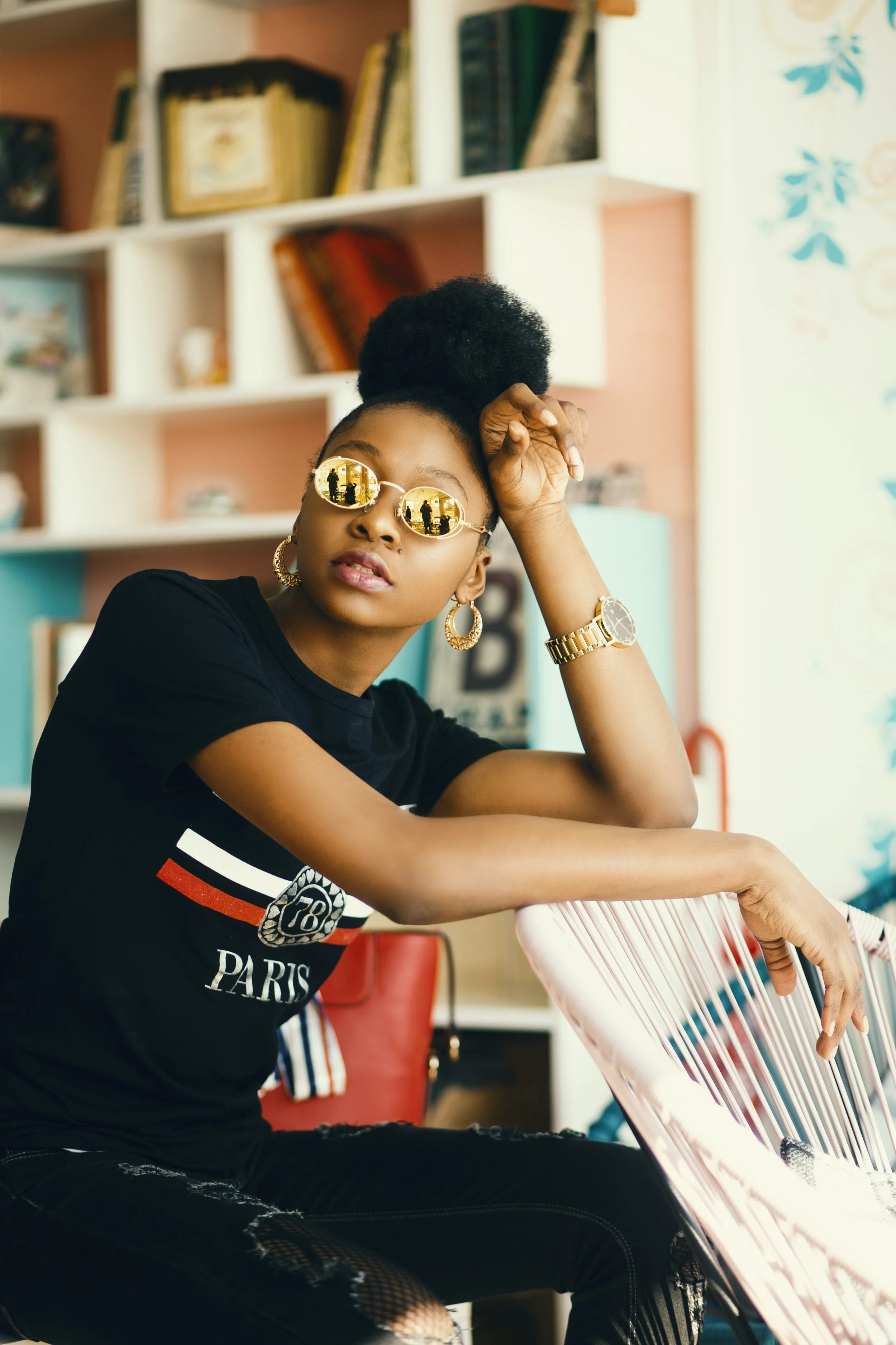 A young woman with dark skin wearing sunglasses, gold jewelry, a black t-shirt with a Paris print, and ripped black jeans, sitting indoors with a colorful background, resting her arm on a chair.
