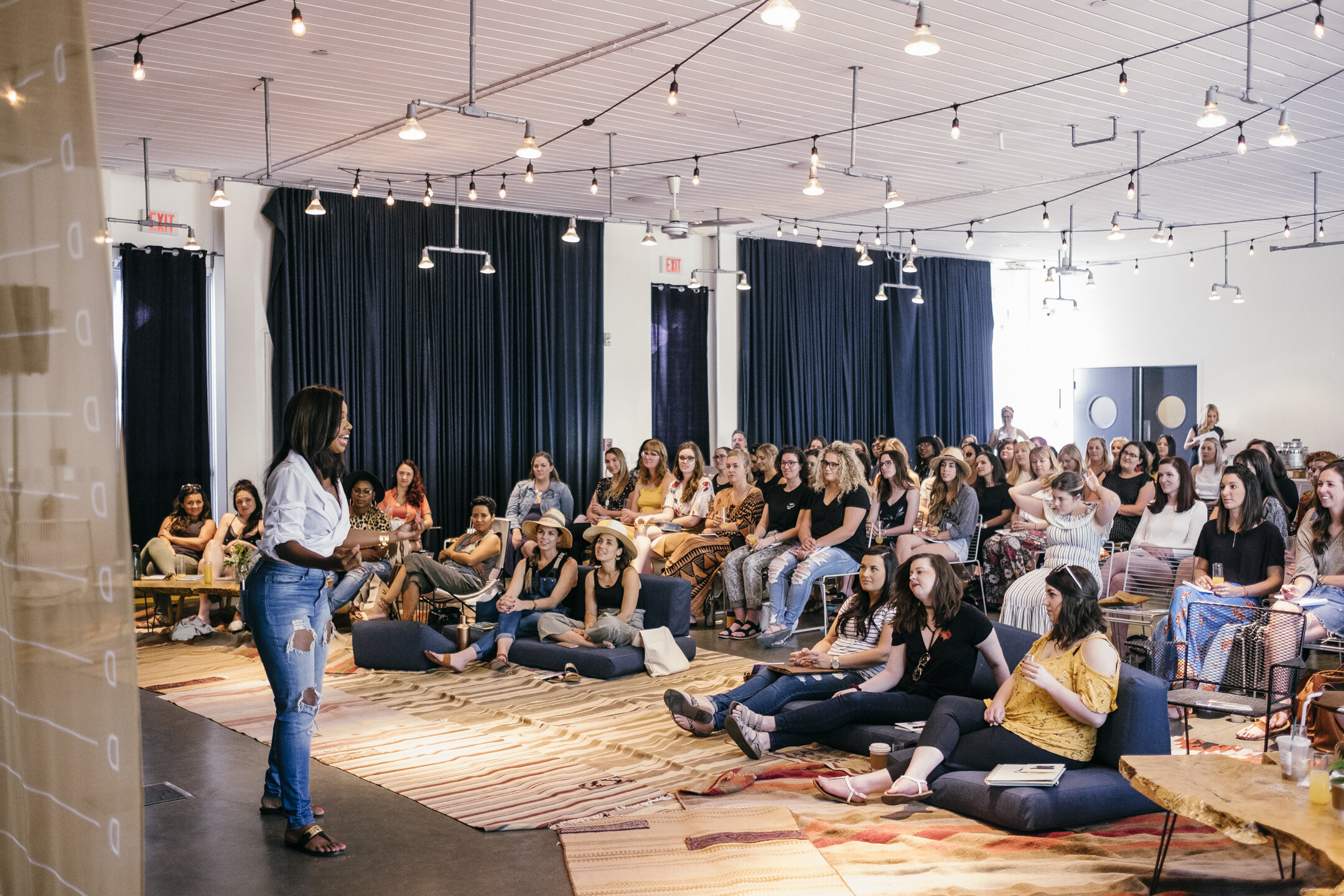 A woman is speaking to an audience in a spacious room with hanging string lights and dark curtains, seated on the floor and chairs, listening attentively.