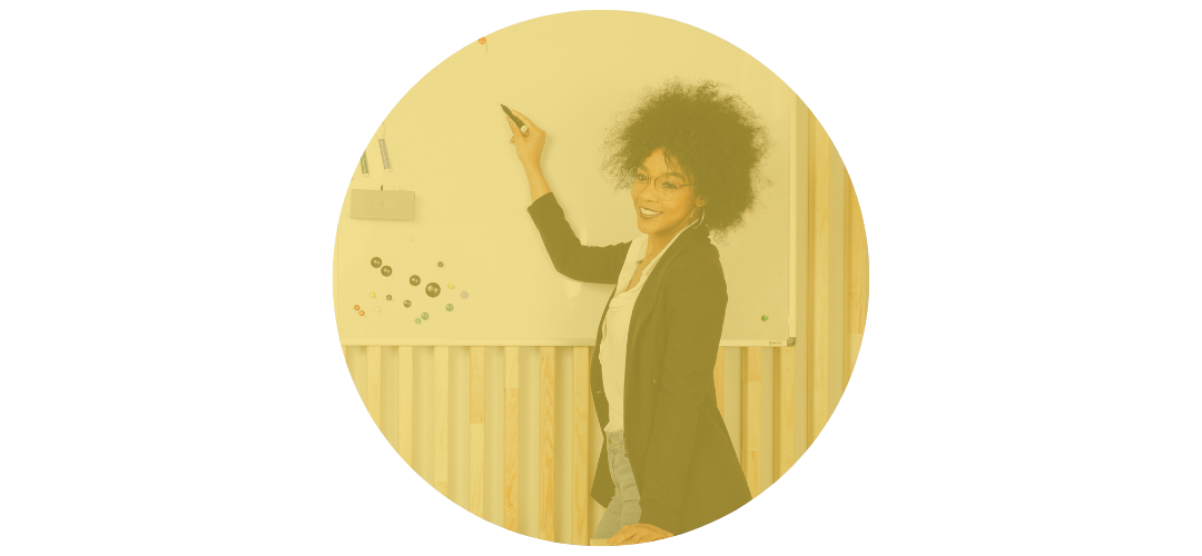 A woman with curly hair and glasses teaching in a classroom, writing on a whiteboard.