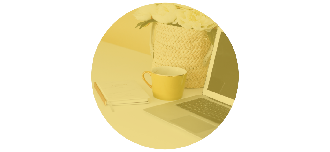 A white desk with a yellow mug, notebook, and a laptop, with a flower arrangement in a woven basket in the background.
