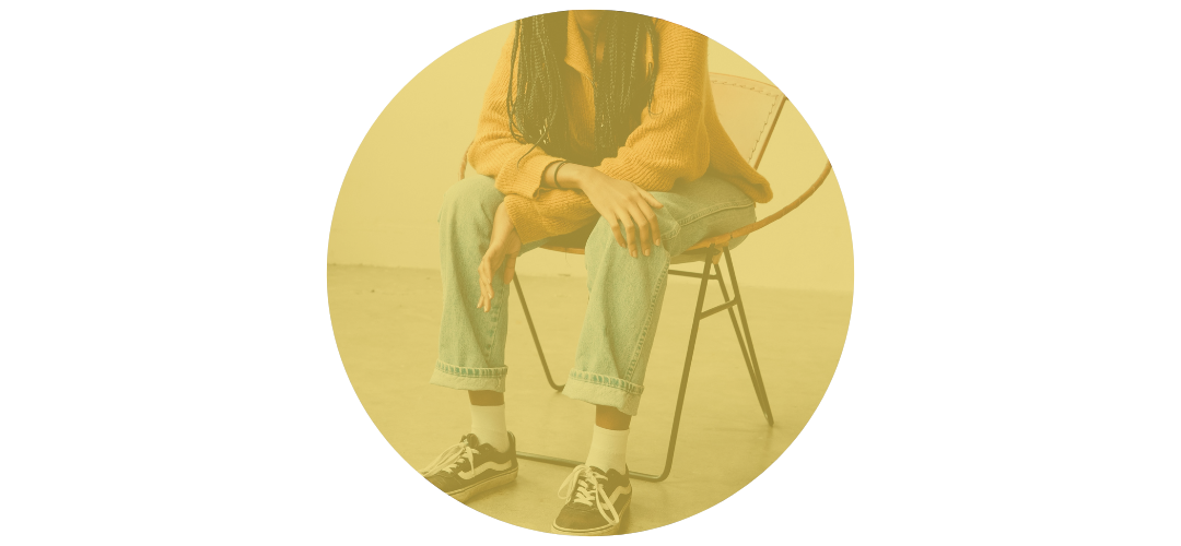Person sitting on a chair with crossed legs, wearing jeans, sneakers, a yellow sweater, and a black shirt underneath, in an indoor setting.