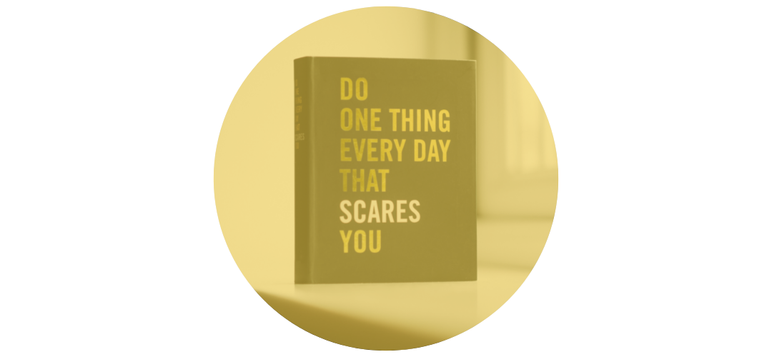 A book with the title 'Do One Thing Every Day That Scares You' is standing upright on a flat surface. The background appears to be a neutral-colored wall and a blurred object, possibly a mug, is present to the right of the book.