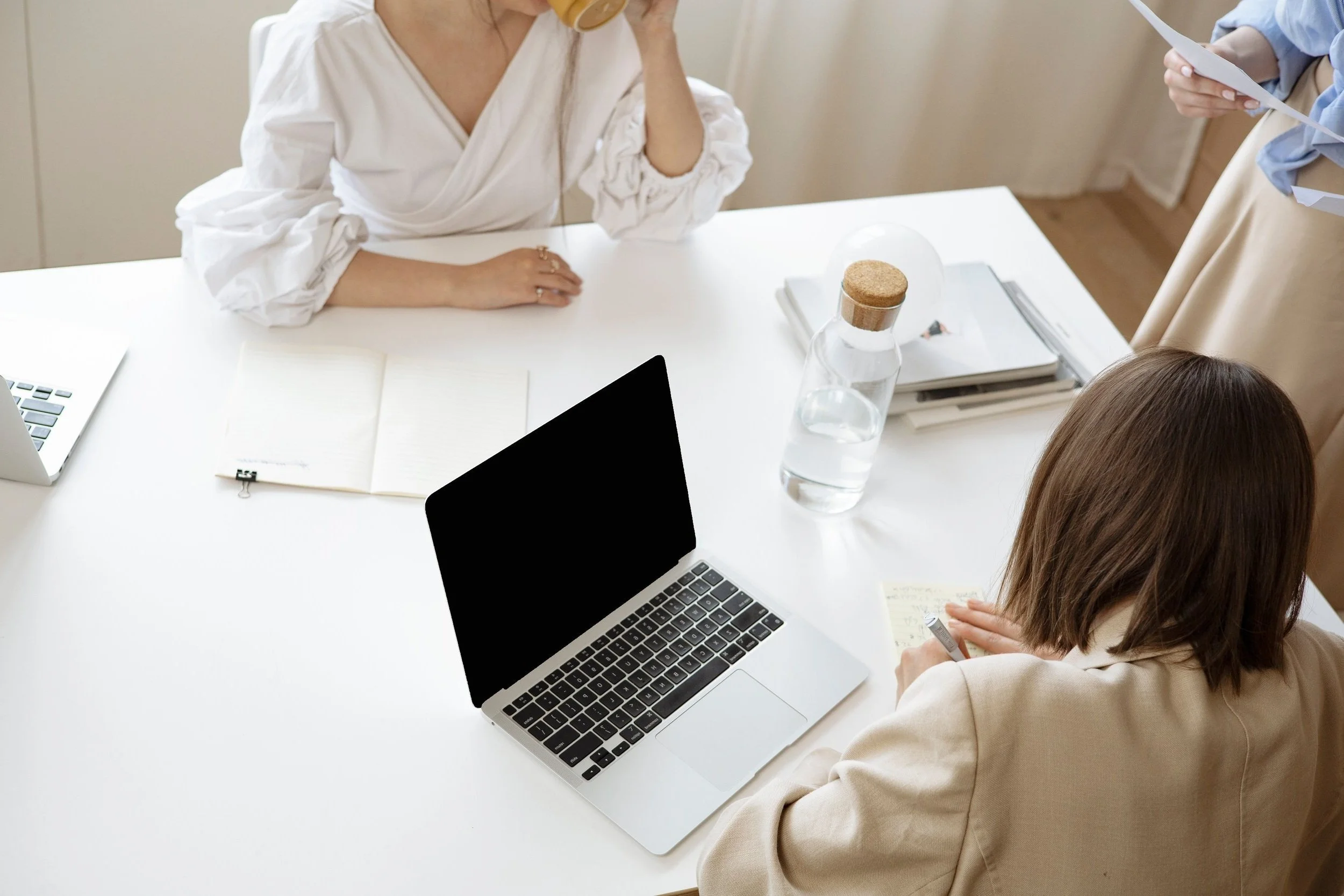 Three women in a meeting around a white table with laptops, notebooks, a glass water bottle, and papers.