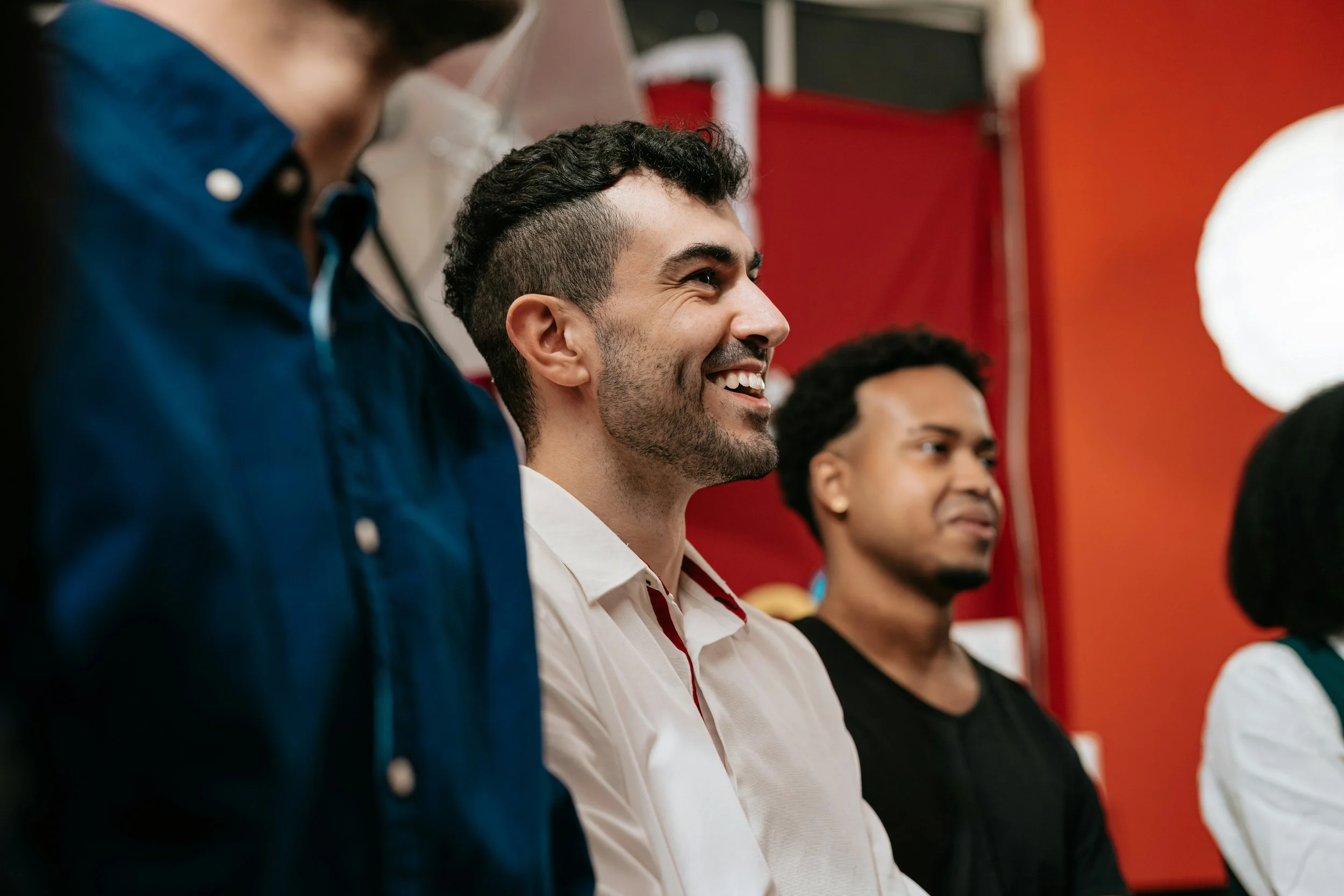 Three men sitting side by side in a room with red walls, smiling and listening attentively, possibly at a presentation or event.