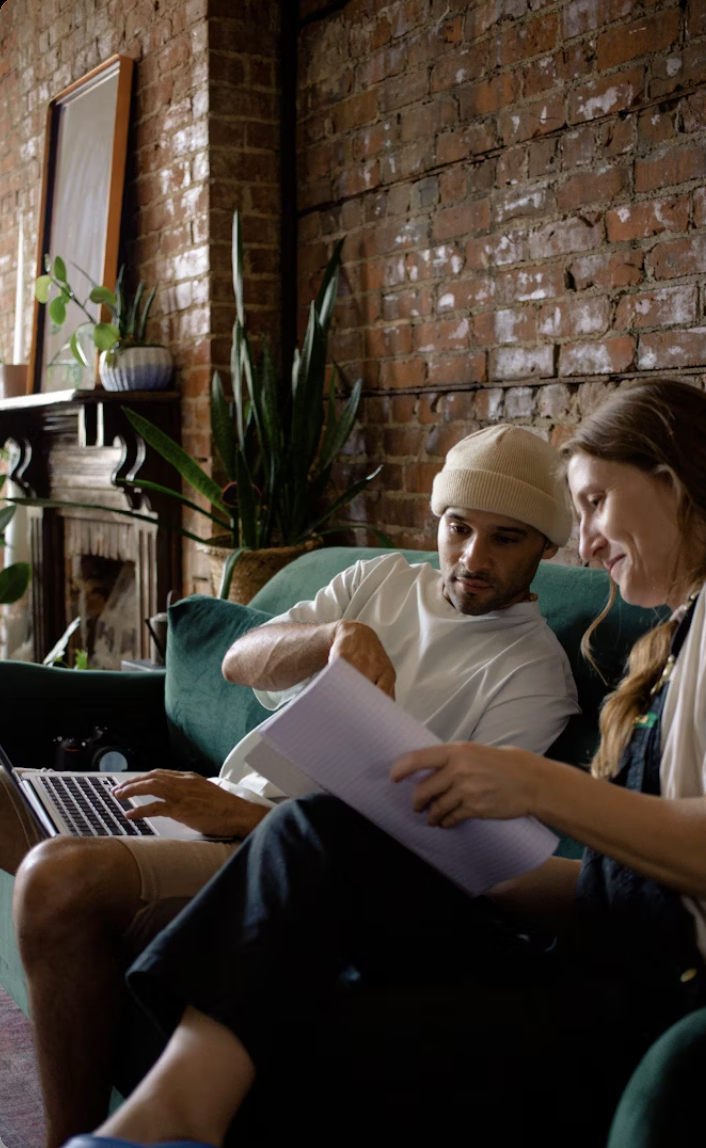 Two people sitting on a green couch, one wearing a beige beanie and white shirt, and the other with long hair, reviewing papers together in a room with brick walls, plants, and a fireplace.