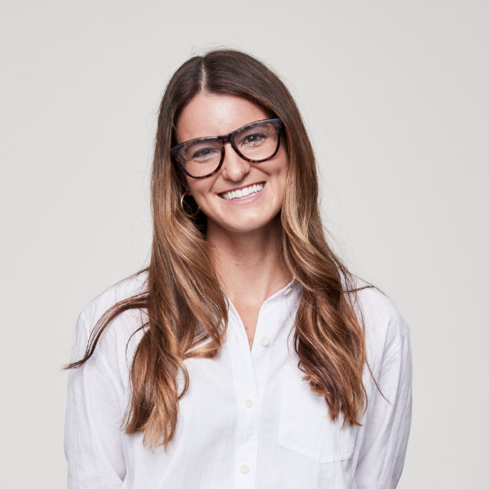 A woman with long wavy brown hair, wearing glasses, a white button-up shirt, and smiling.