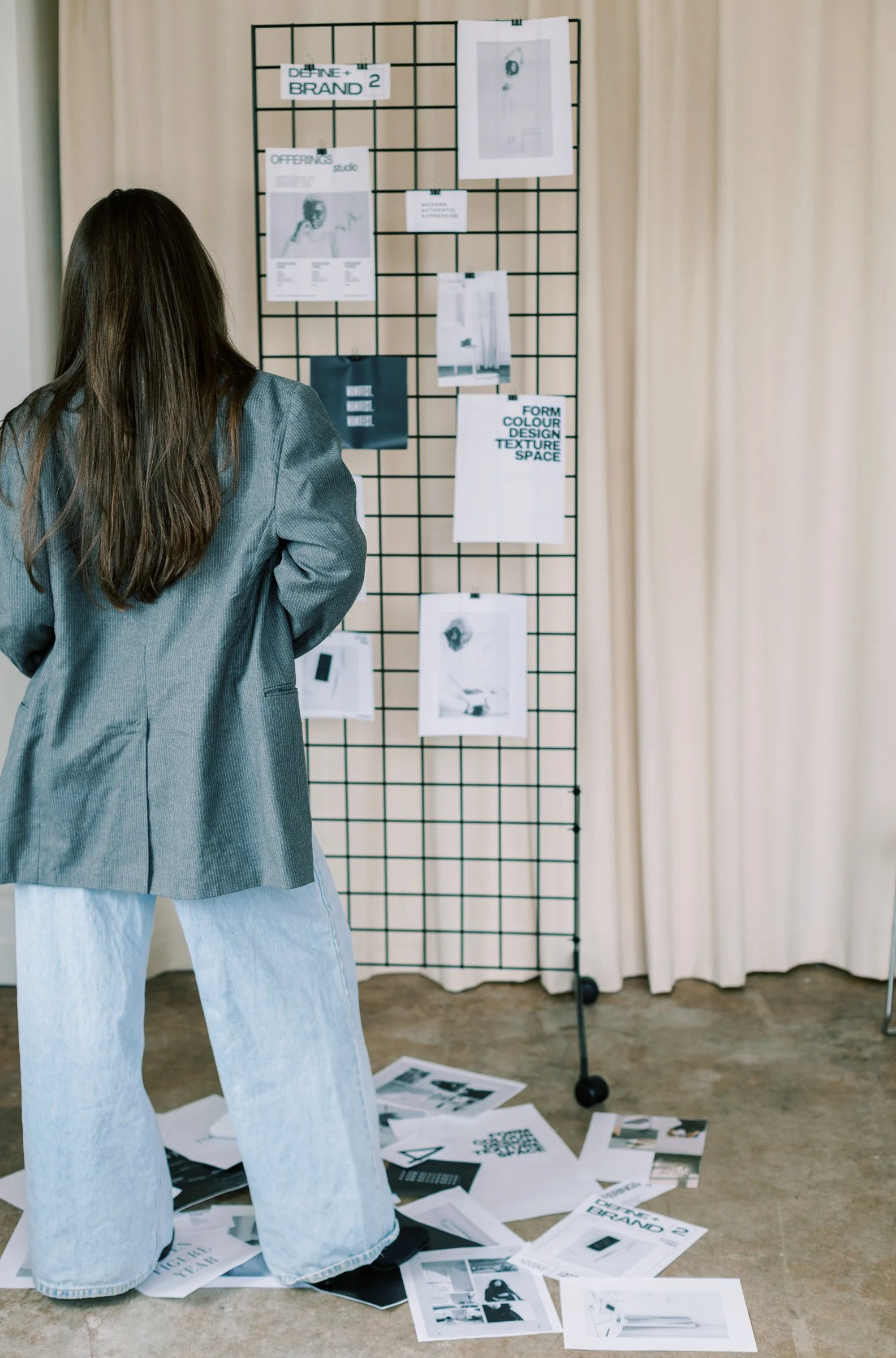 A woman with long hair, wearing a gray blazer and light-washed jeans, stands floor-side facing a black grid display with various papers and images attached to it in an indoor setting.