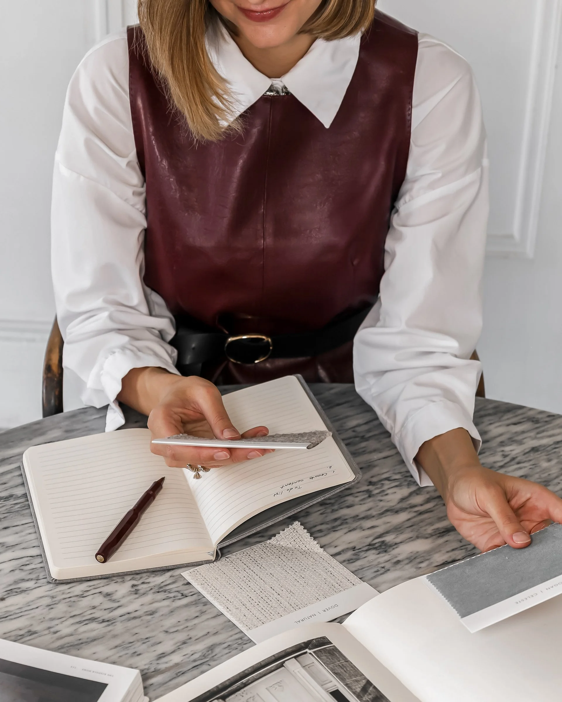 A woman sitting at a marble table, holding a sample of fabric in one hand and pointing to an open notebook with the other. The notebook has a pen on it. Various fabric samples and magazine pages are spread on the table.