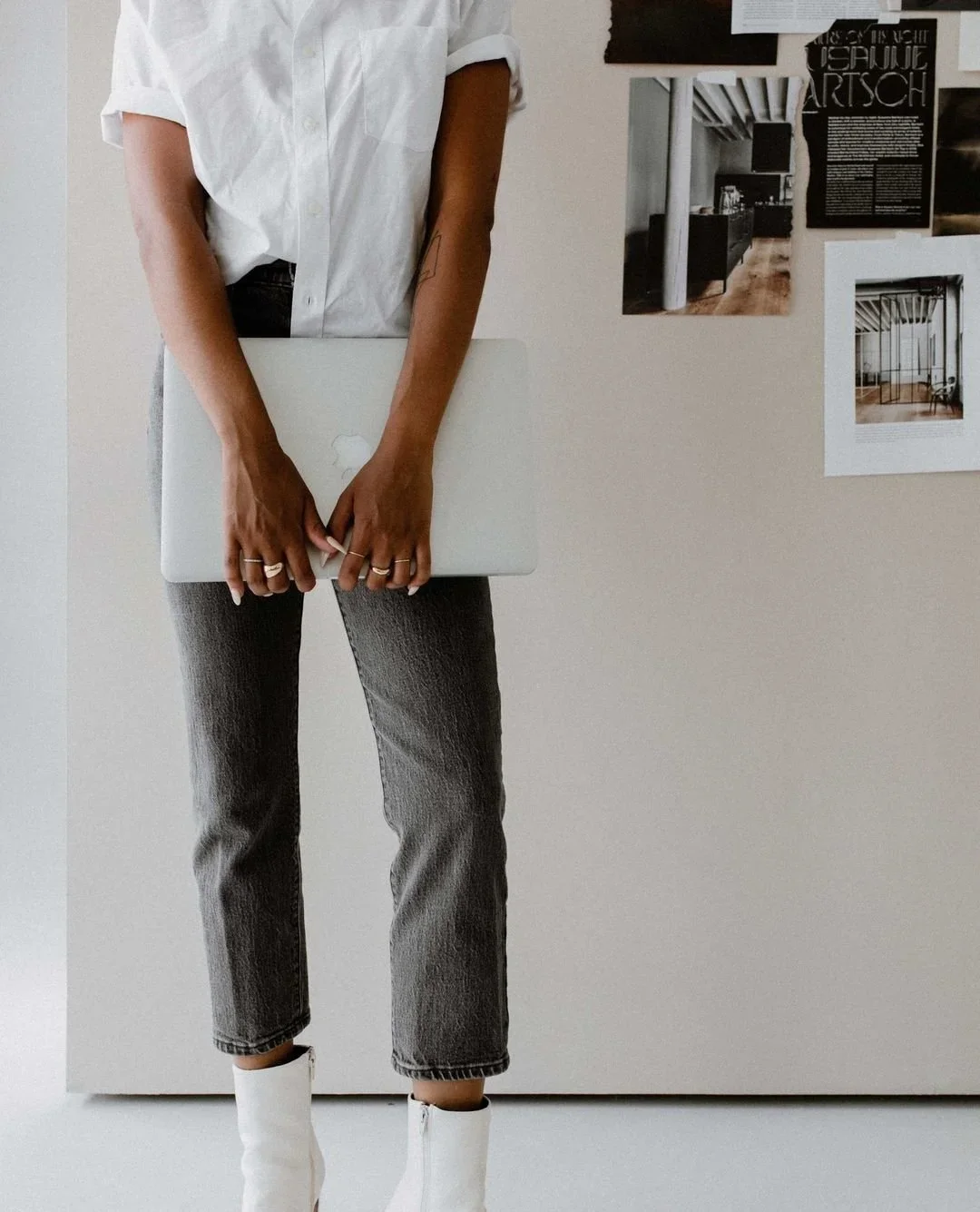 Person holding a closed silver laptop, standing against a plain white wall with black and white art posters.