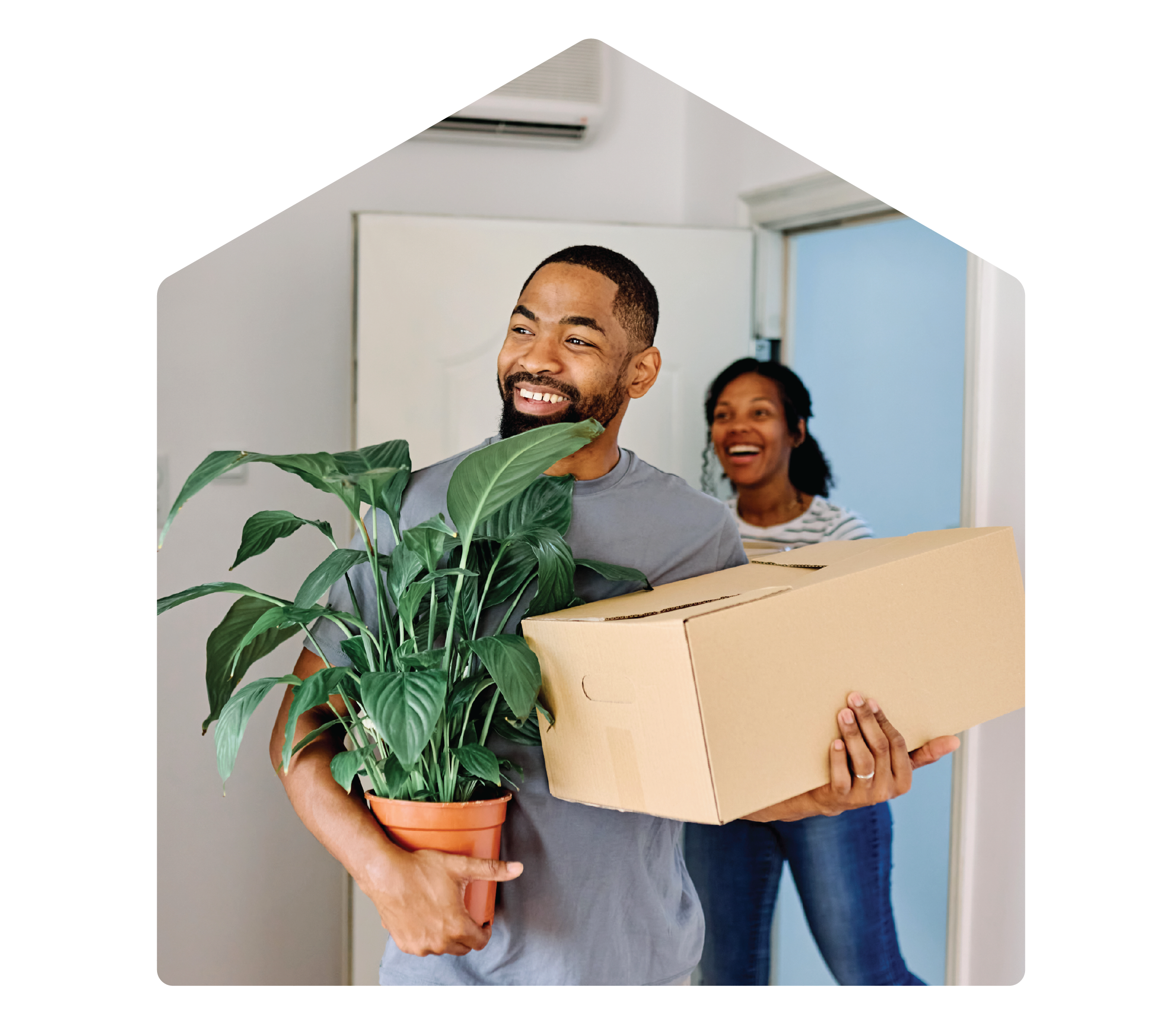 Man carrying a plant and box into his new home. His wife follows behind and is in the doorway.