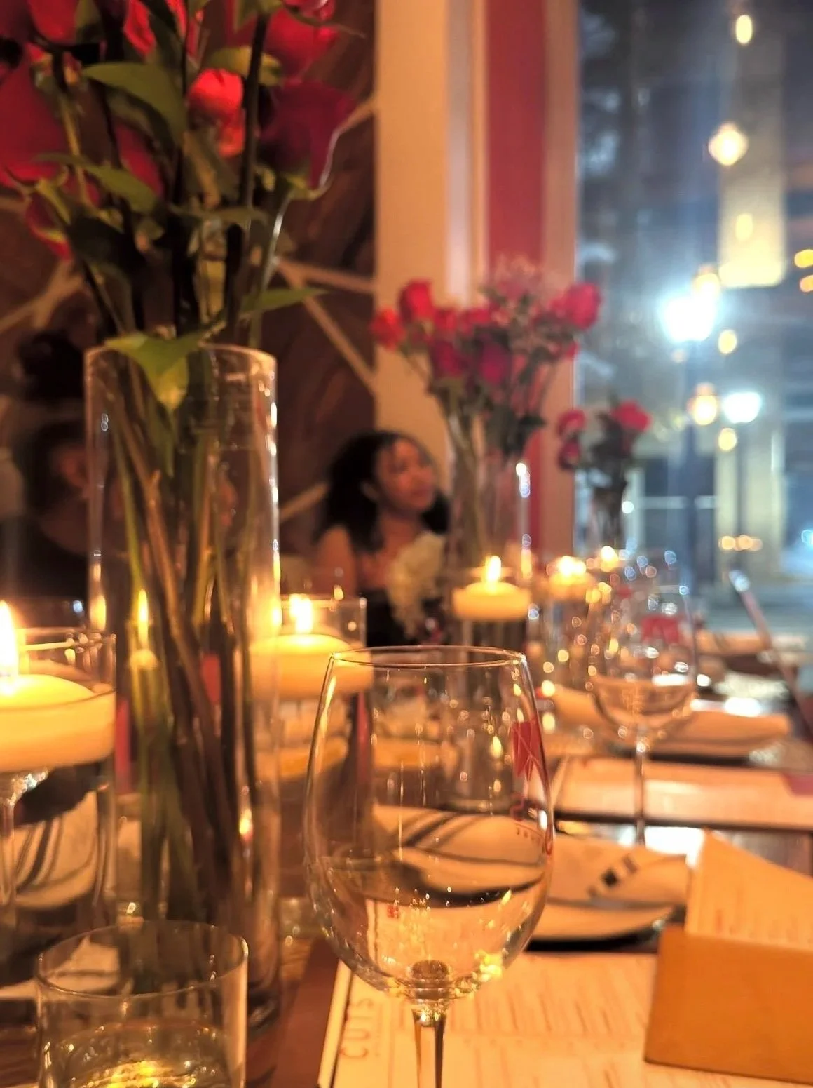A cozy dining table with lit candles, glasses, and floral arrangements inside a restaurant, with a woman sitting in the background near a window showing city lights.