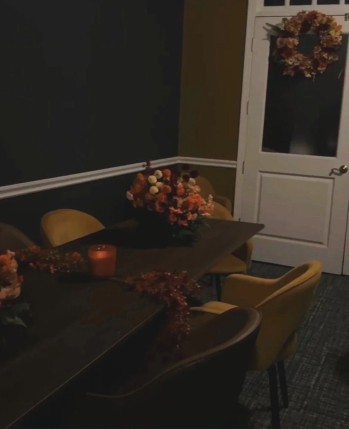 A dining room with a dark wooden table decorated with orange and peach flower arrangements and a lit candle. Yellow upholstered chairs are positioned around the table. There is a white door with a glass panel and a wreath hanging on it, and dark walls with white trim.