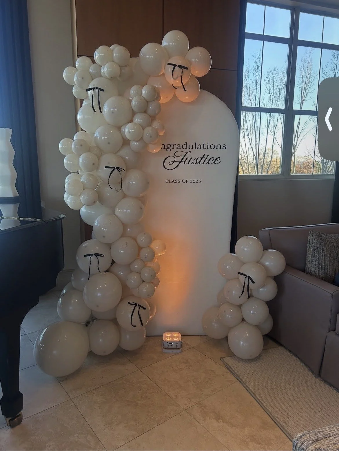 White balloon display with small black ribbon bows surrounding a curved backdrop that reads "congratulations Justice CLASS OF 2025"; the balloons are arranged to form a crescent shape on either side of the backdrop with some climbing over it, and a light illuminates from the bottom.