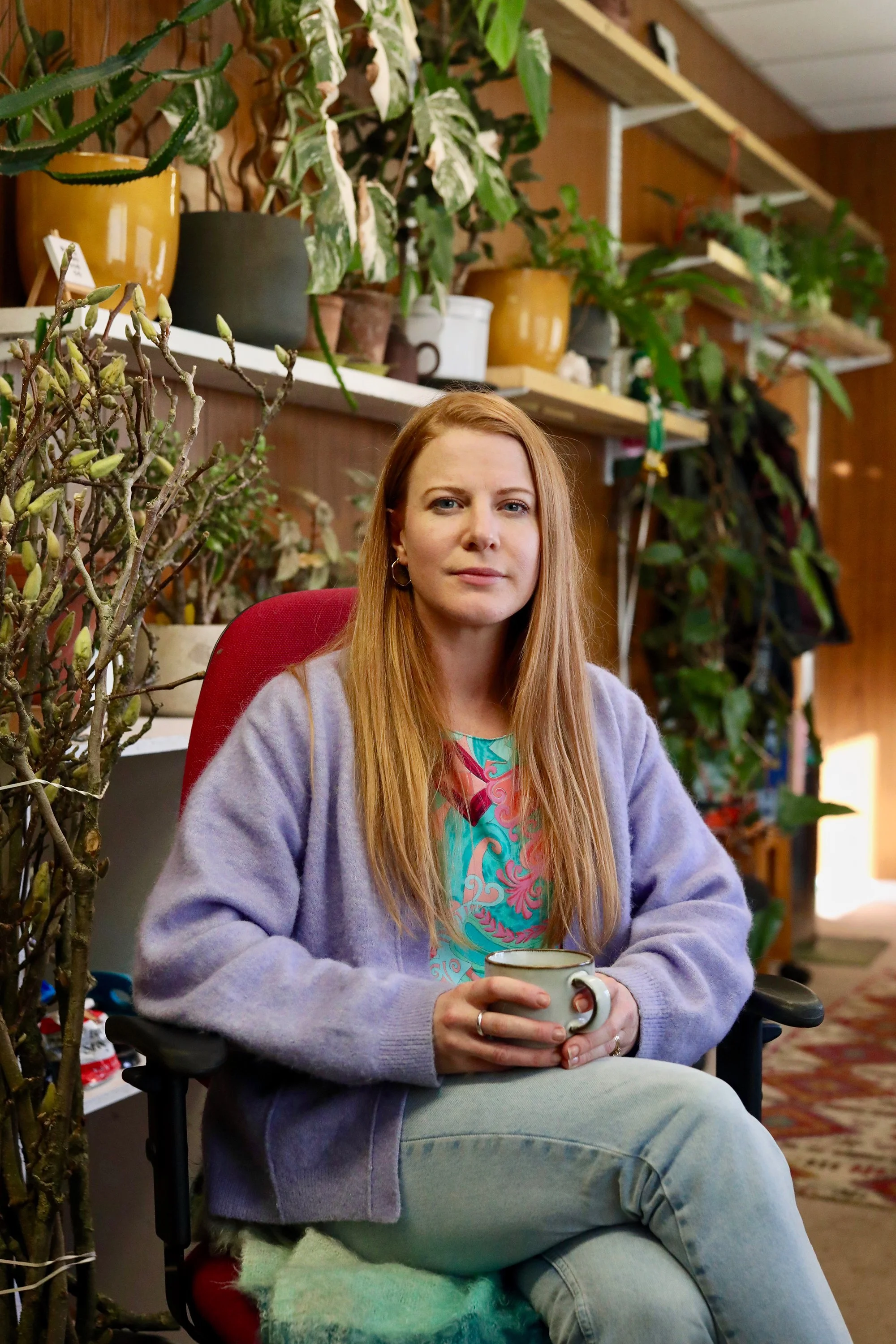 A woman with long red hair sitting in a red chair, holding a mug, in a room filled with plants and wooden shelving.