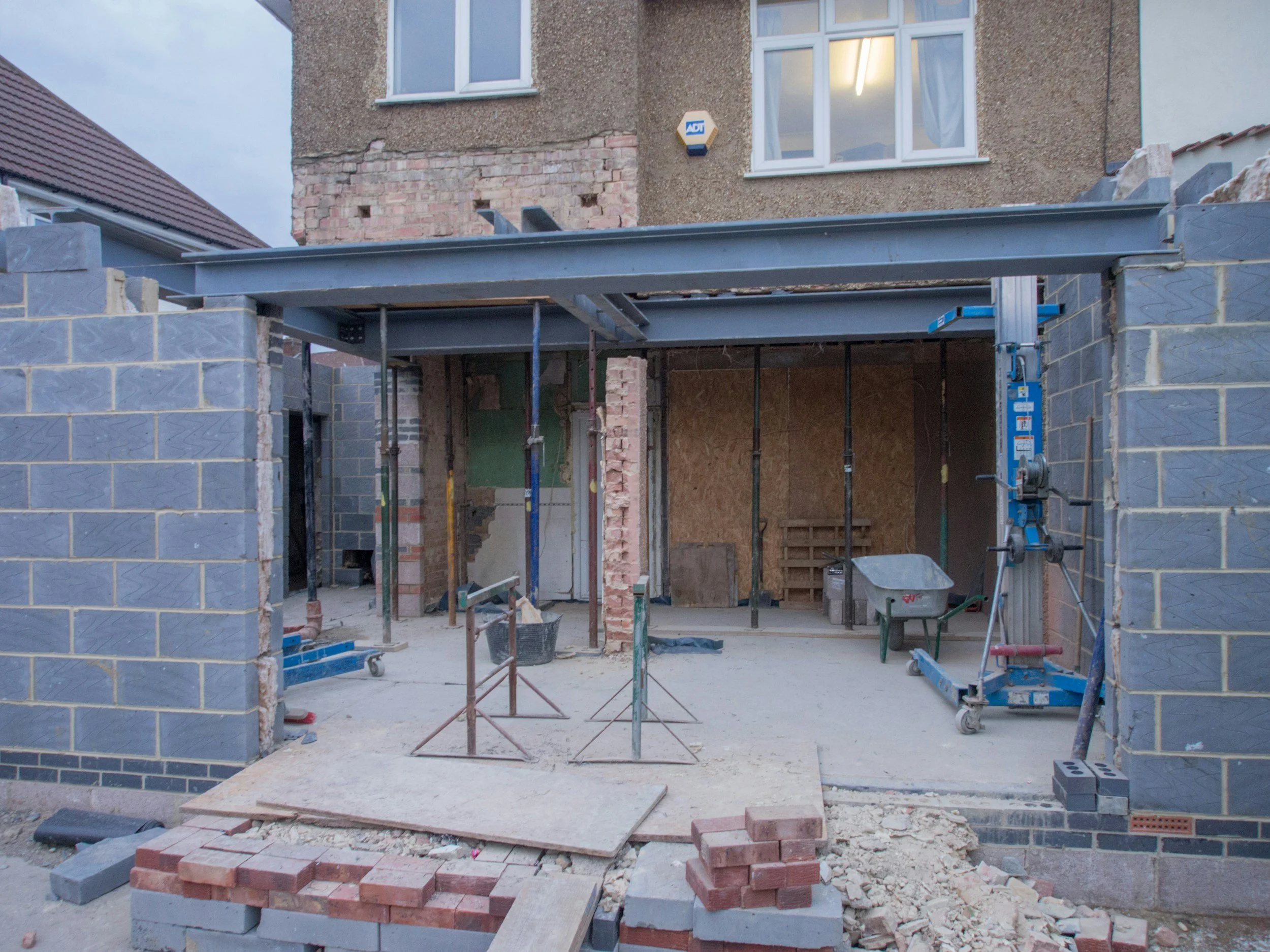 Construction site of a residential extension, showing scaffolding, bricks, and construction tools inside a partially built structure with walls and an opening for a door or window.