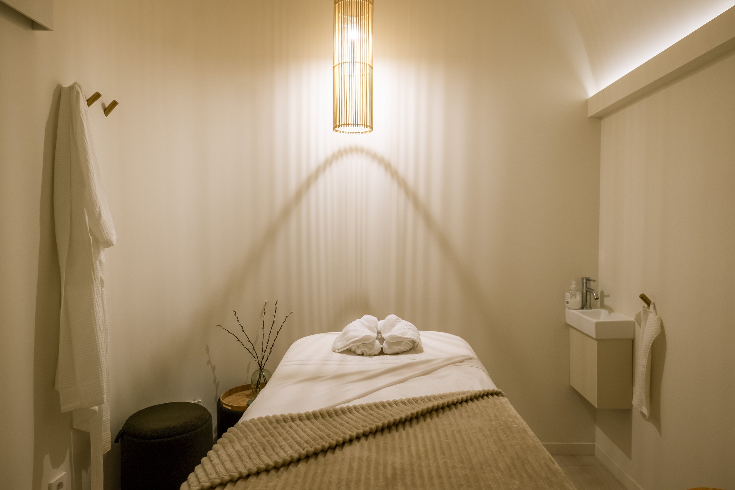 Sala de masaje minimalista, limpia con camilla beige, toallas dobladas, lámpara colgante de madera, pared blanca, con sombra geométrica de la lampara con luz tenue cálida.